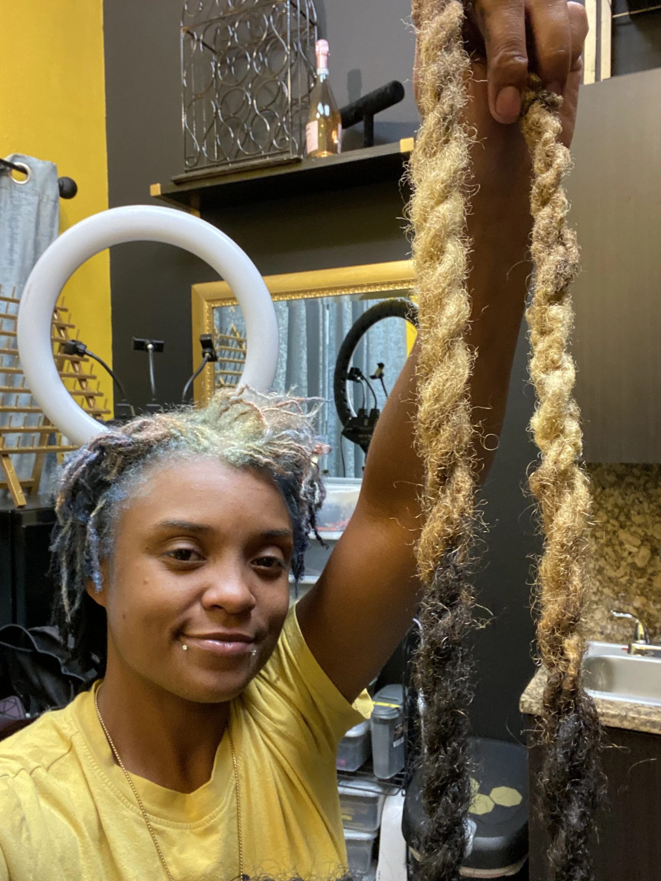 A woman with multicolored dreadlocks, smiling and holding up two long locks of her hair in a room with mirrors, lighting equipment, and kitchen cabinets in the background.