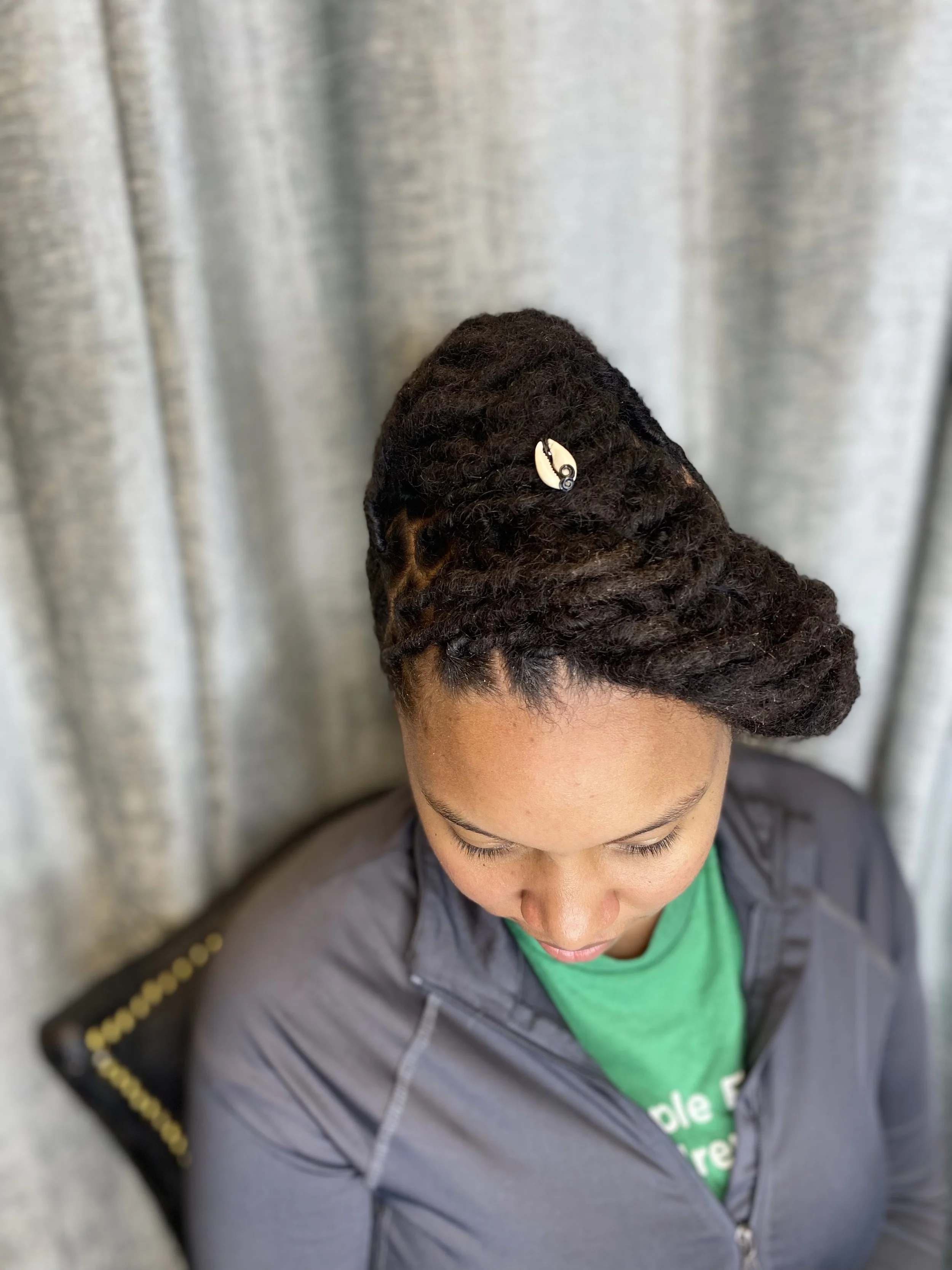 Woman with styled dreadlocks sitting against a silver curtain, wearing a gray jacket with black and gold detailing and a green shirt underneath.