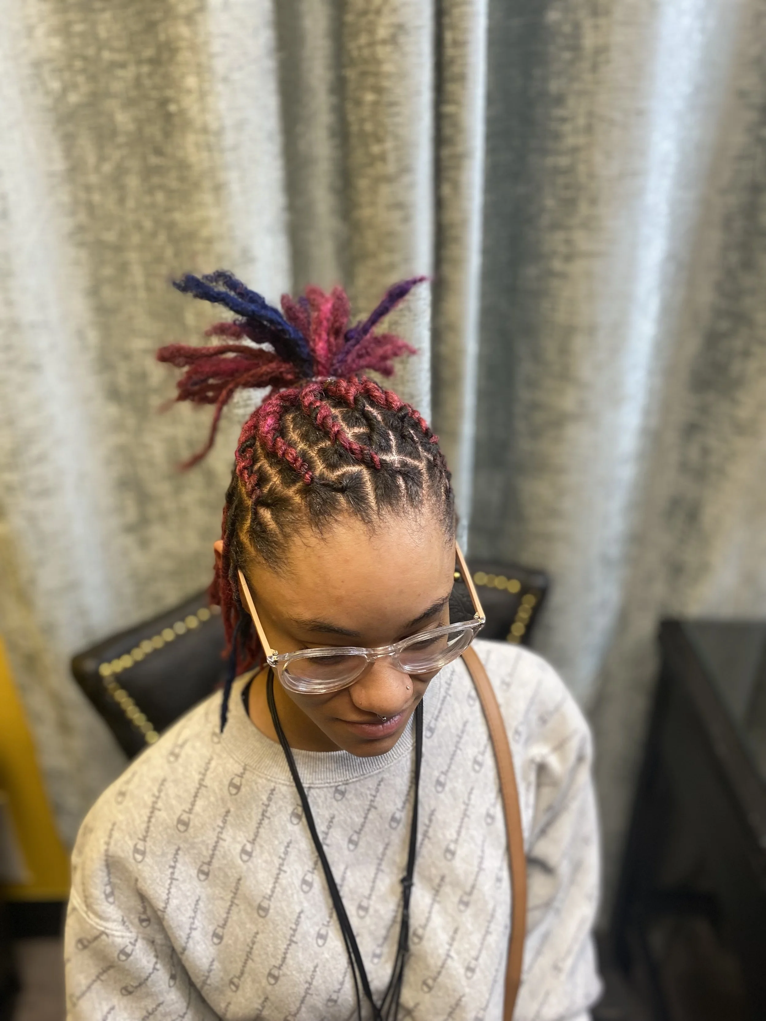 A woman with colorful braided dreadlocks styled in a high bun, including shades of pink and purple, wearing glasses, a gray Champion sweatshirt, and a black lanyard, sitting in front of a metallic curtain.