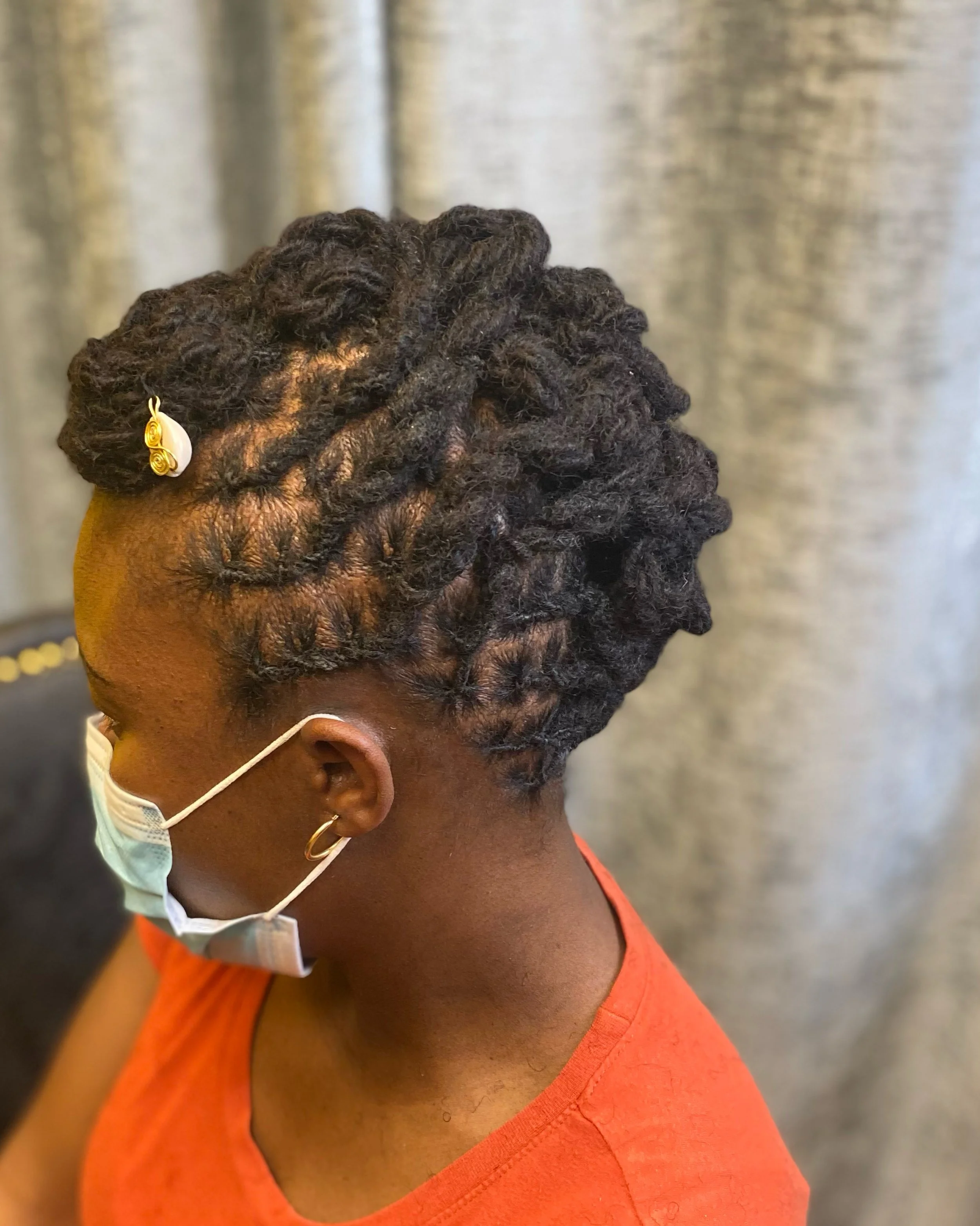 Close-up of a woman with styled dreadlocks adorned with a white decorative hairpin, wearing a face mask, gold earrings, and an orange shirt.