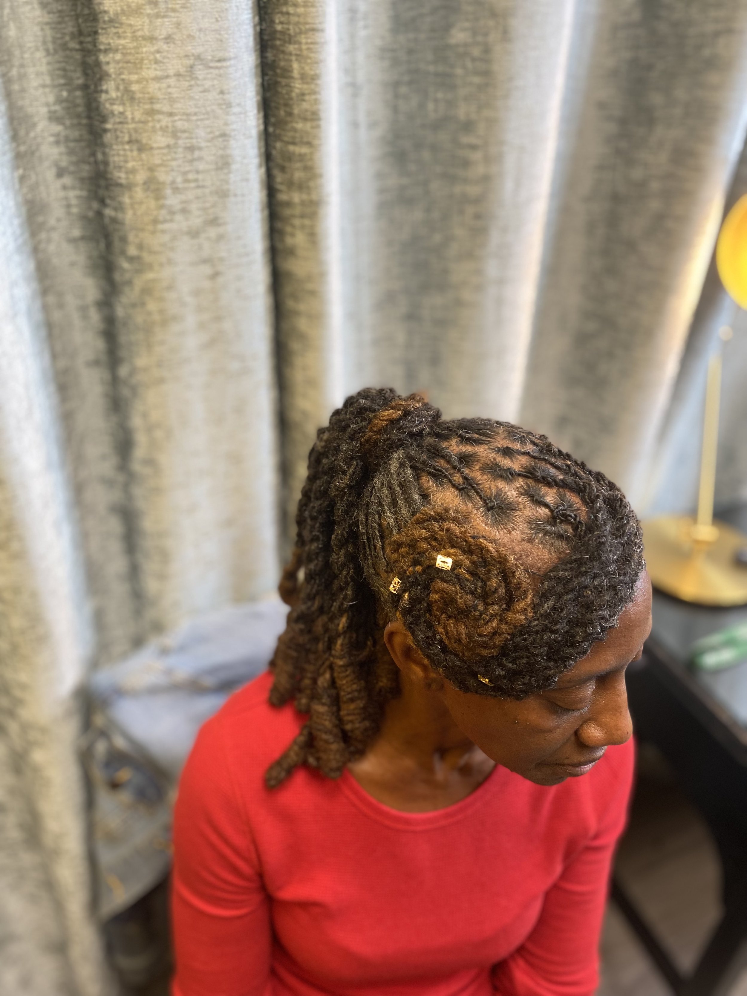 A woman with styled dreadlocks wearing a red top, sitting in front of light-colored curtains.