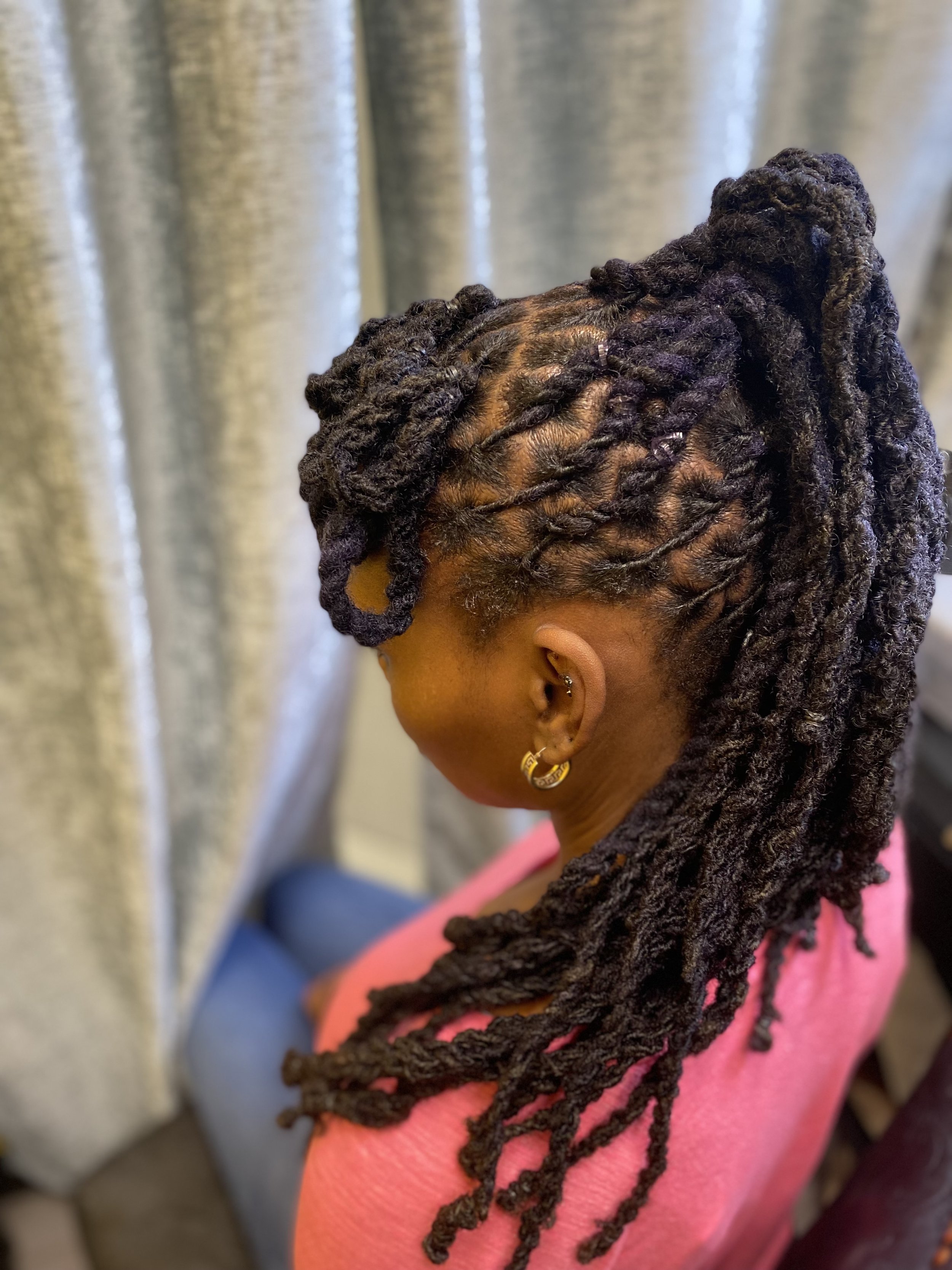 A woman with dark brown, long dreadlocks styled into a high ponytail, sitting indoors with a silver curtain in the background, wearing a pink top and gold hoop earrings.