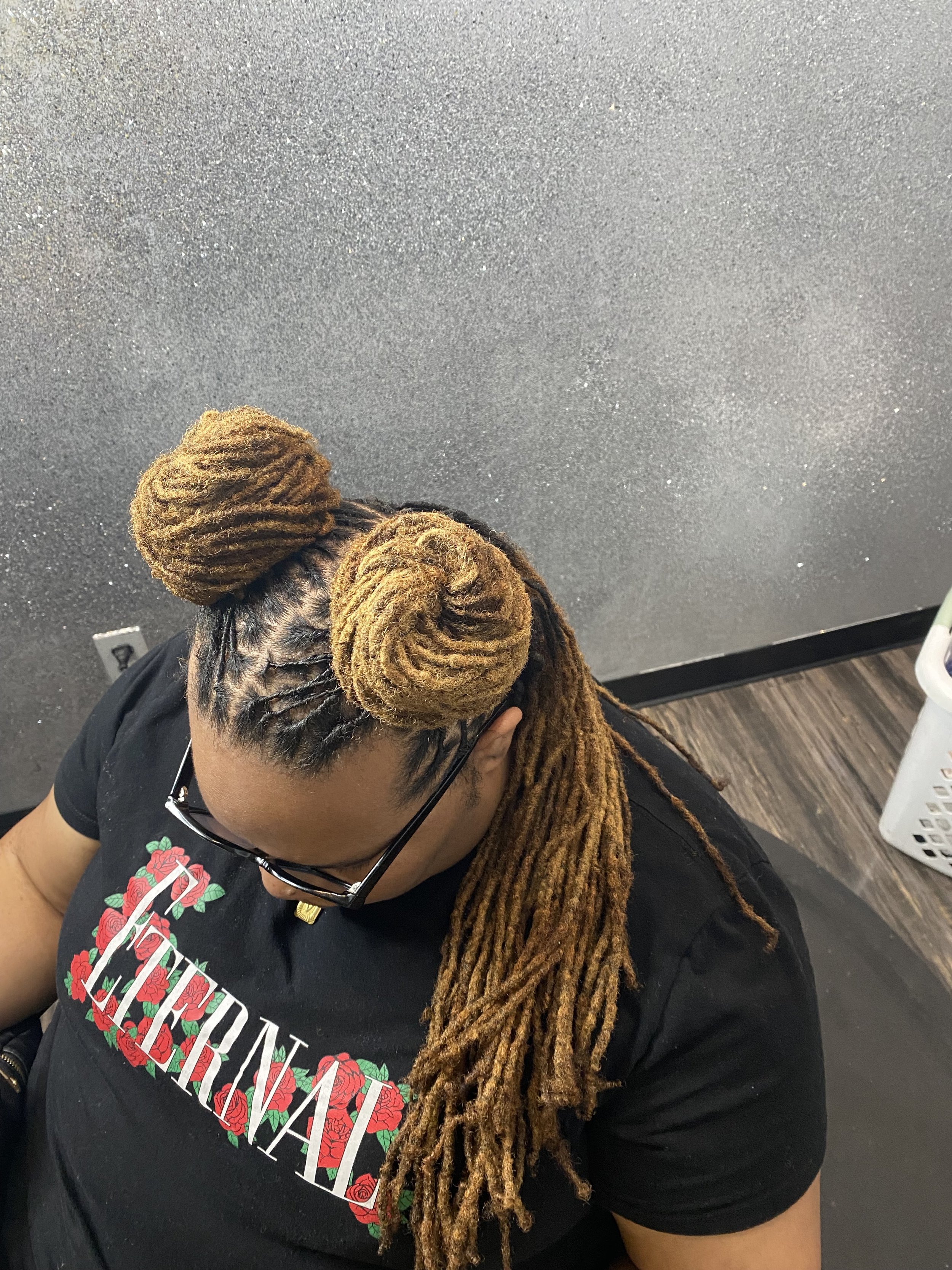 Woman with styled dreadlocks and two large buns on top, wearing glasses and a black T-shirt with red roses and the word 'FIERA' on it, sitting in a room with a sparkly gray wall and dark wooden floor.