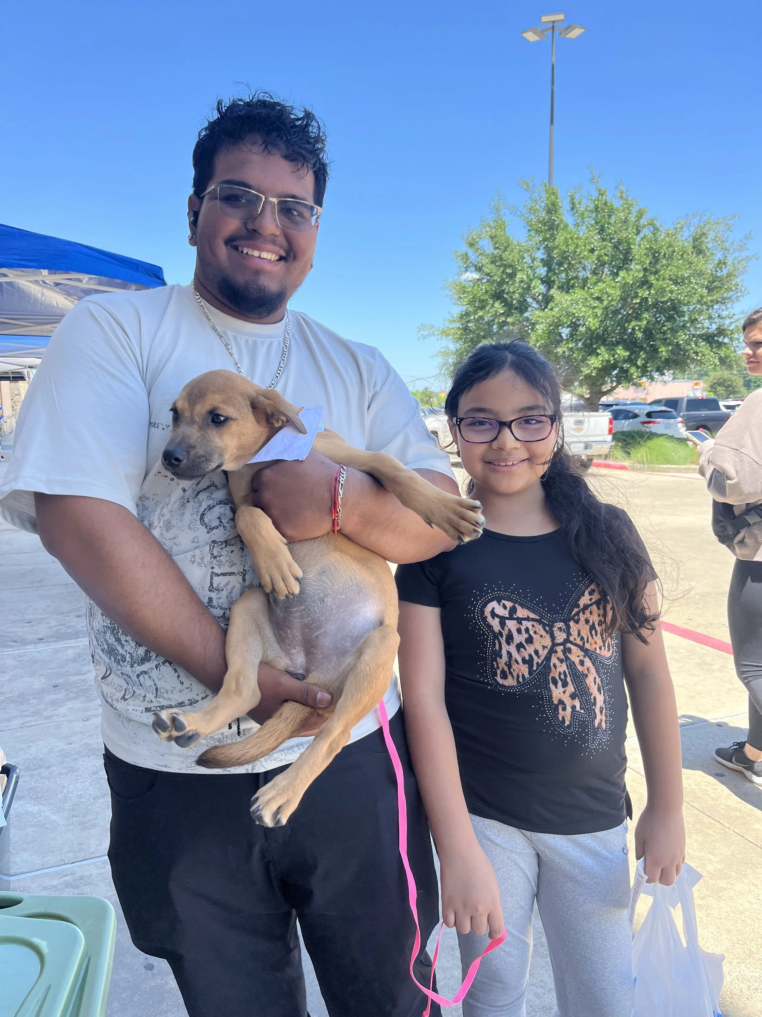A man holding a puppy and a young girl standing outdoors in a parking lot with trees and cars in the background, bright blue sky overhead.