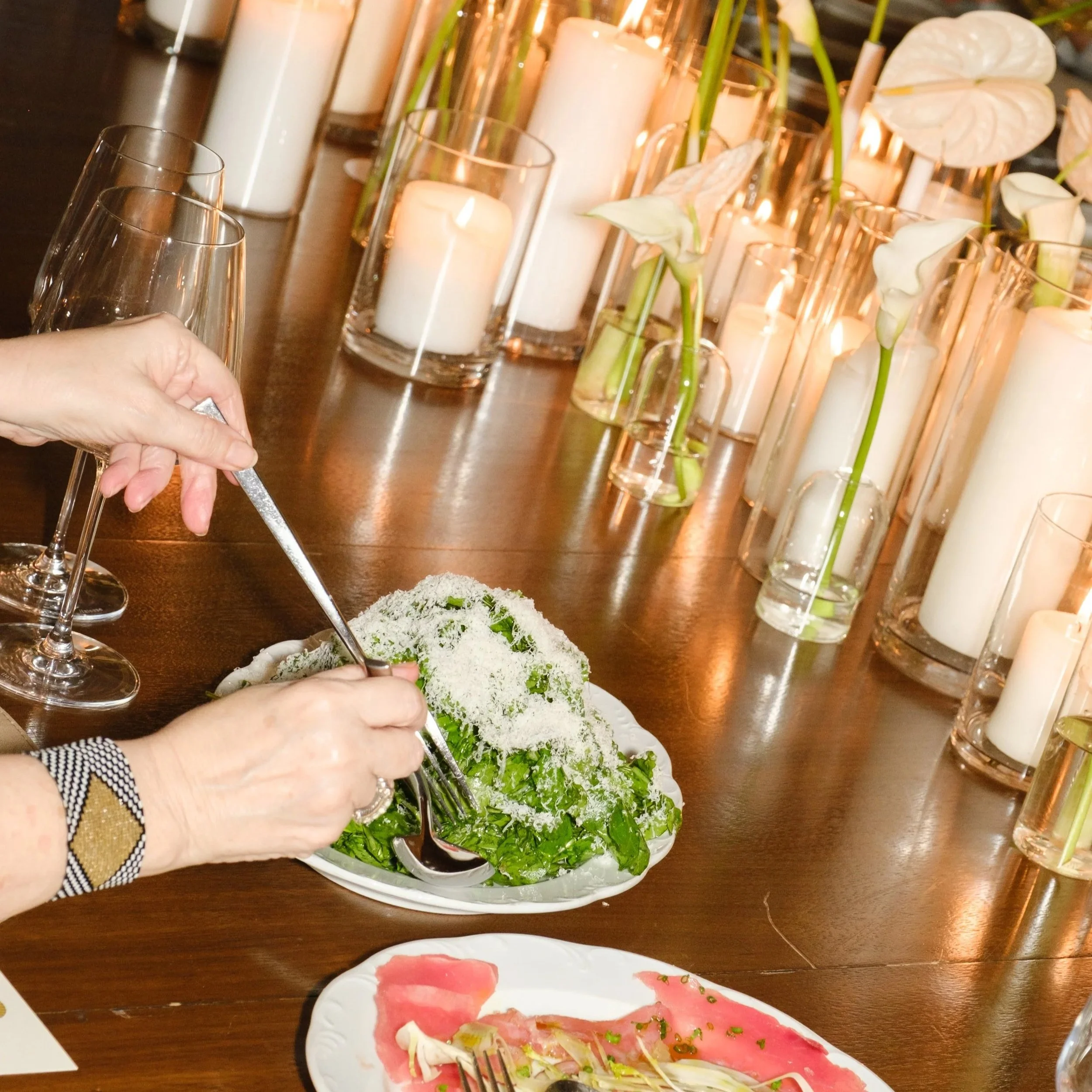 Person placing grated cheese on a salad with greens, on a dining table decorated with candles and white flowers.