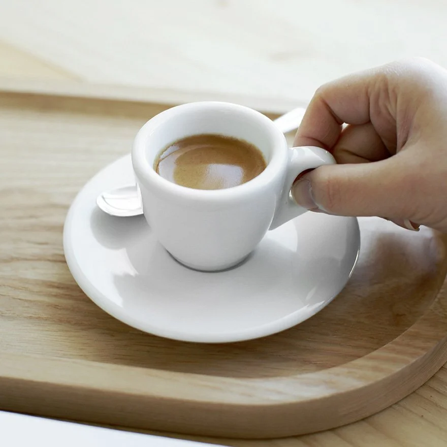 Hand, die eine kleine weiße Espressotasse mit Espresso hält, auf einem weißen Unterteller auf einem runden Holztablett