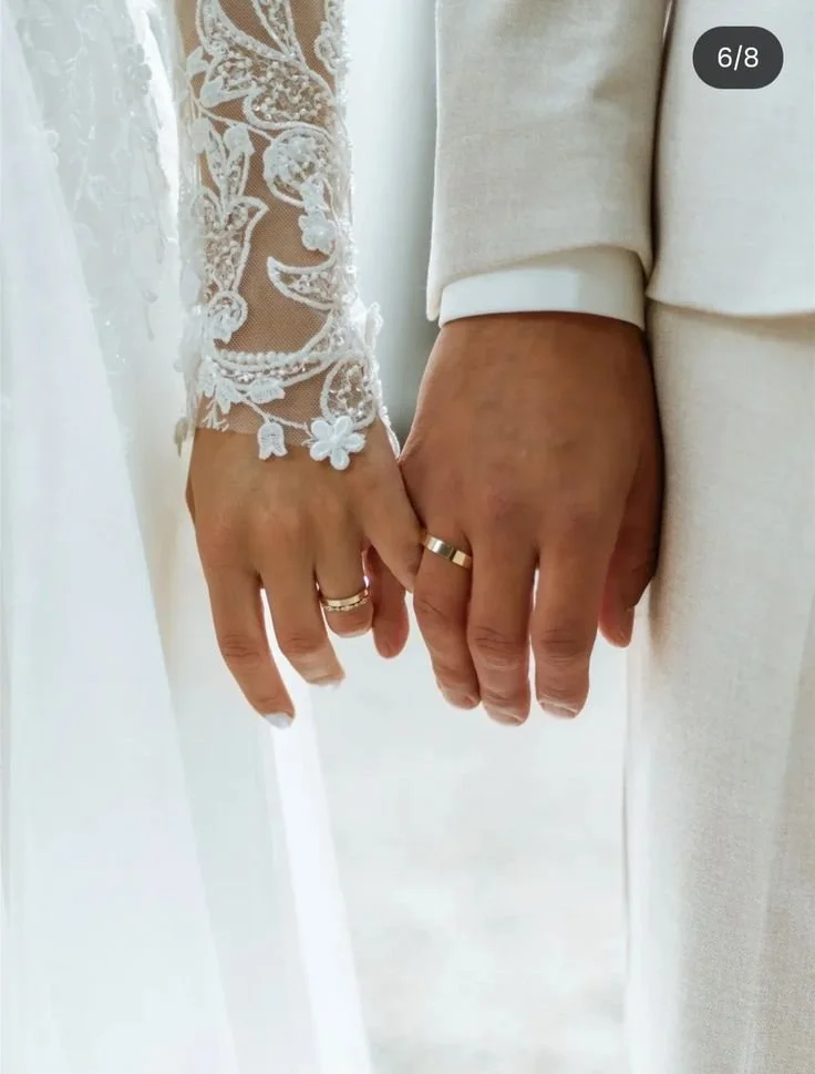 A bride and groom holding hands outdoors, with a rocky landscape in the background. The bride is wearing a white wedding dress with a beaded upper portion, and the groom is in a dark pinstripe suit.