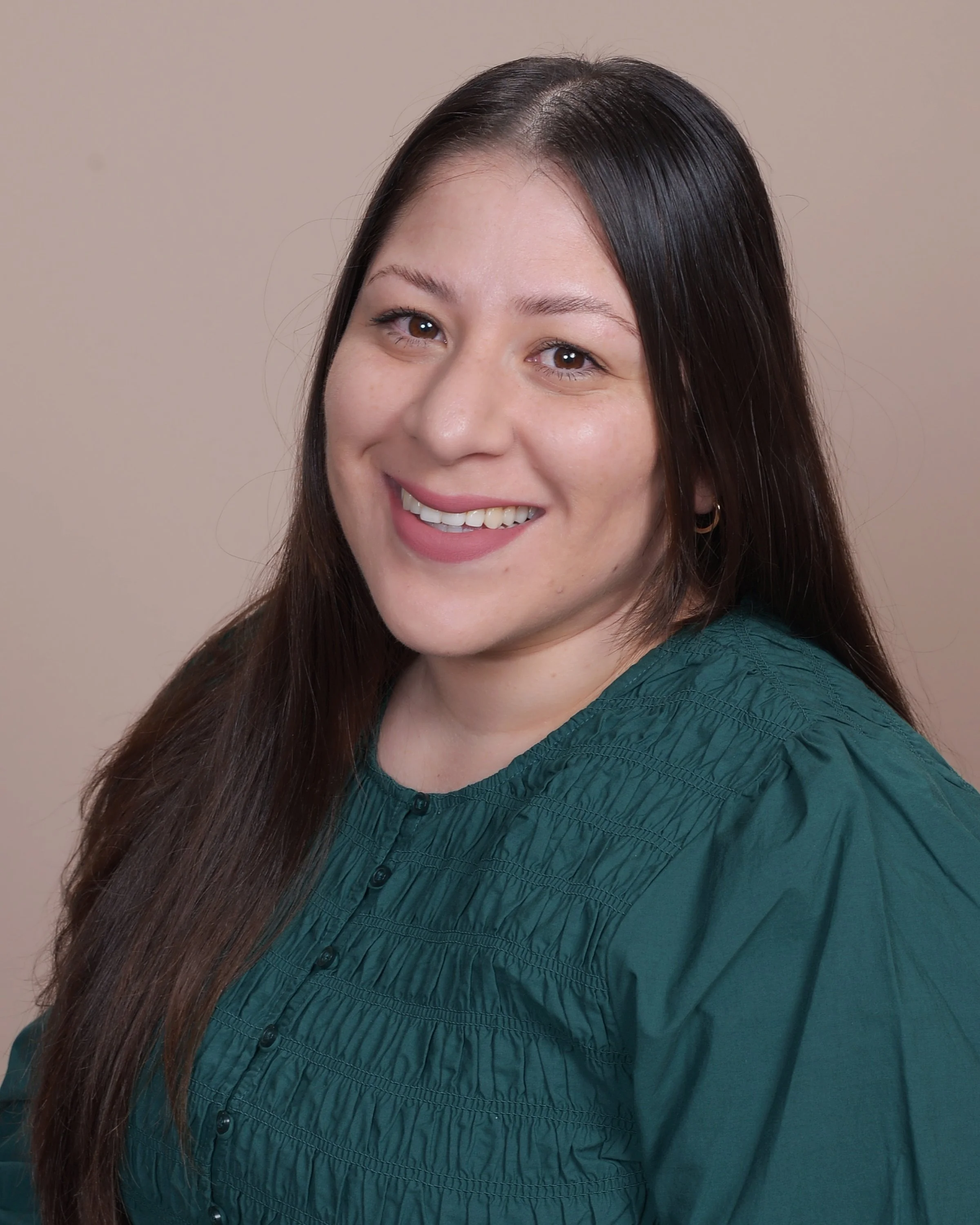 A woman with long brown hair, wearing a pink sleeveless dress, sitting outdoors in front of a historic stone building, smiling at the camera.