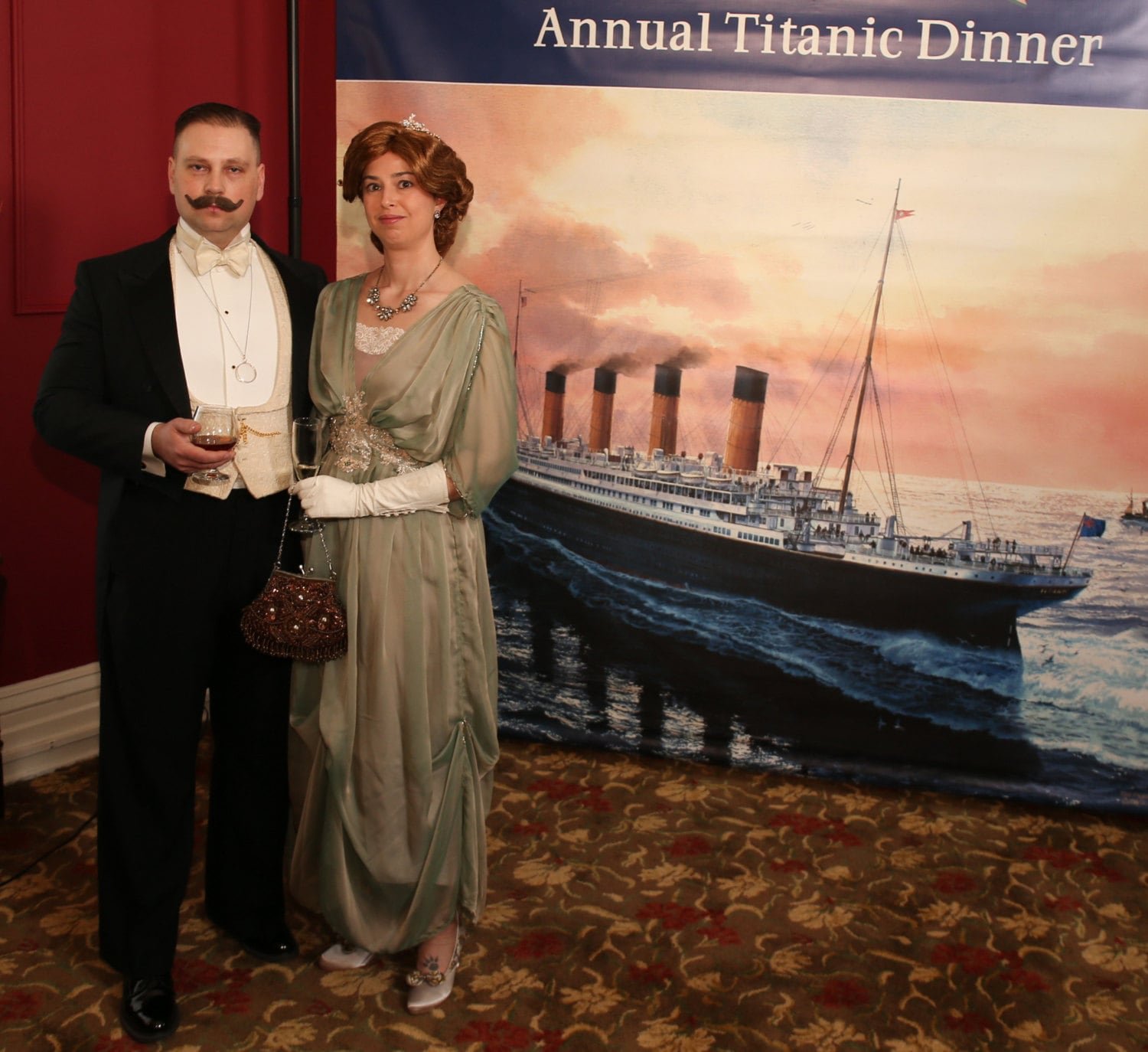 Man and woamn in Edwardian clothes posing in front of the big Titanic poster.