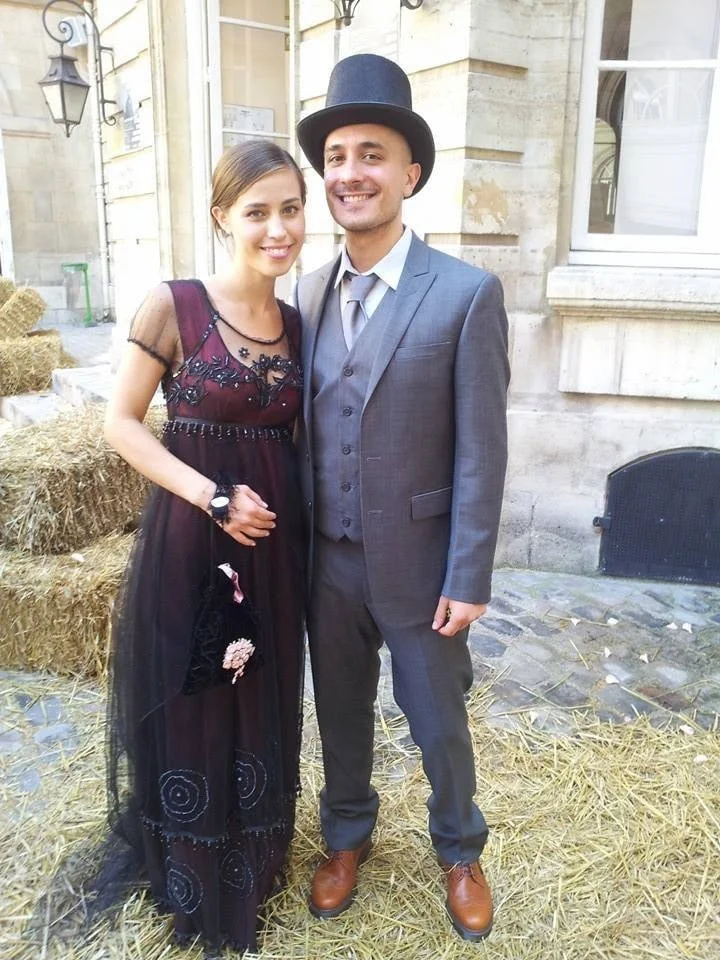 Picture of a couple - young lady in the Titanic jump dress holding a small bag , standing next to a man in a grey suite and a hat.
