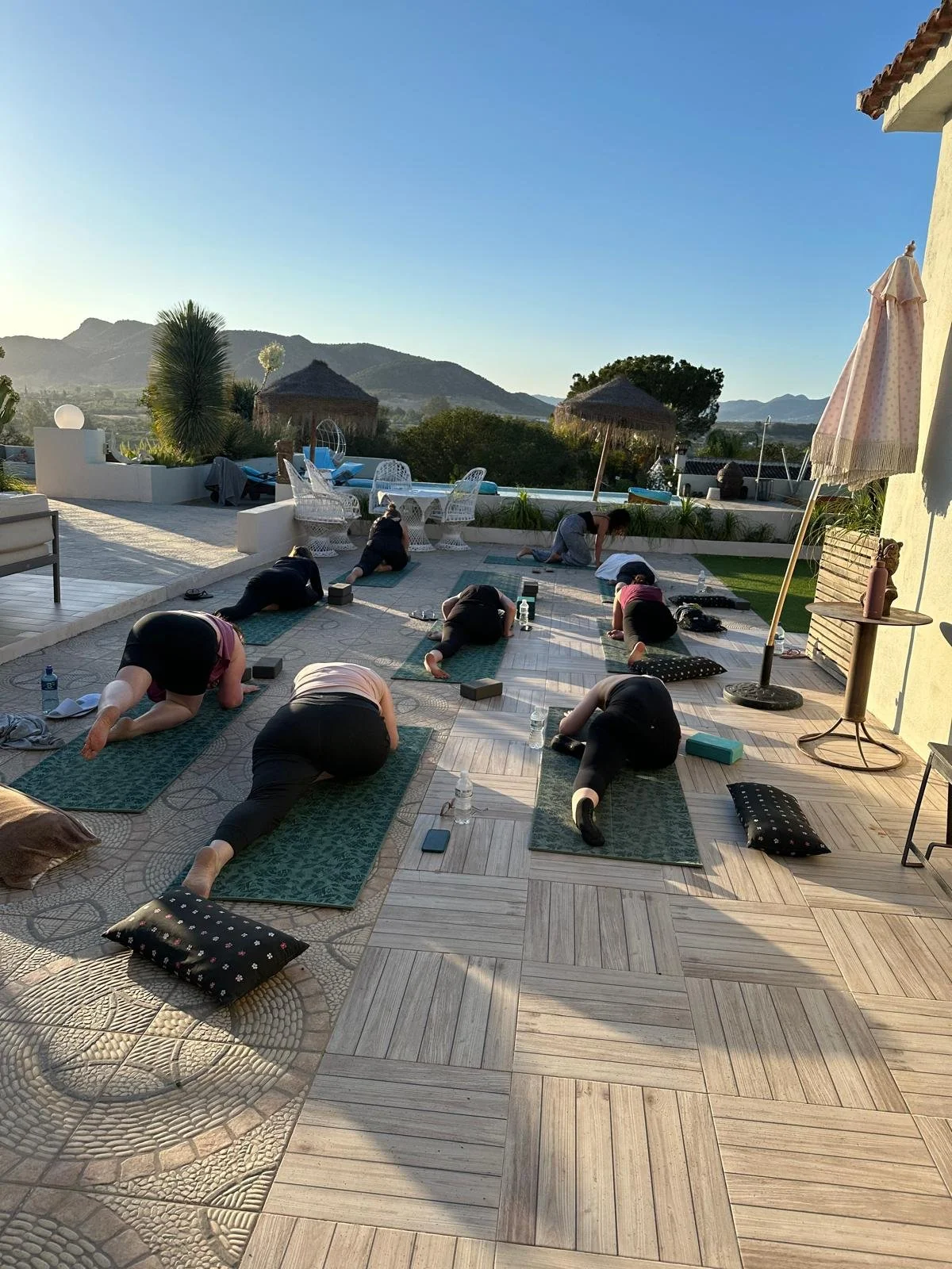 Group of people practicing yoga on mats outdoors with mountains in the background, during a sunny day.