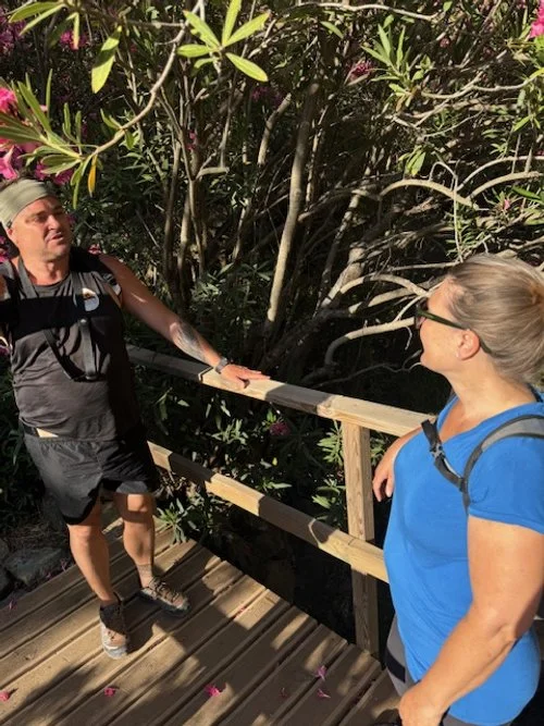 Two people talking on a wooden walkway surrounded by foliage and pink flowers, wearing casual outdoor attire and backpacks.