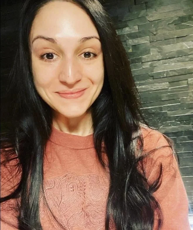 Morgan our yoga teacher, smiling in front of a dark stone wall.