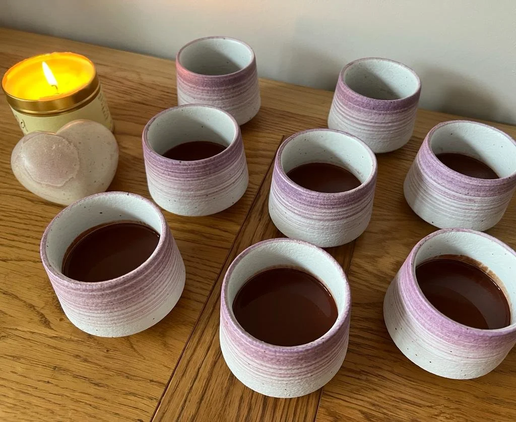 Ceramic cups with purple gradient rims holding chocolate drink, placed on a wooden surface; a lit candle and a heart-shaped stone are nearby.