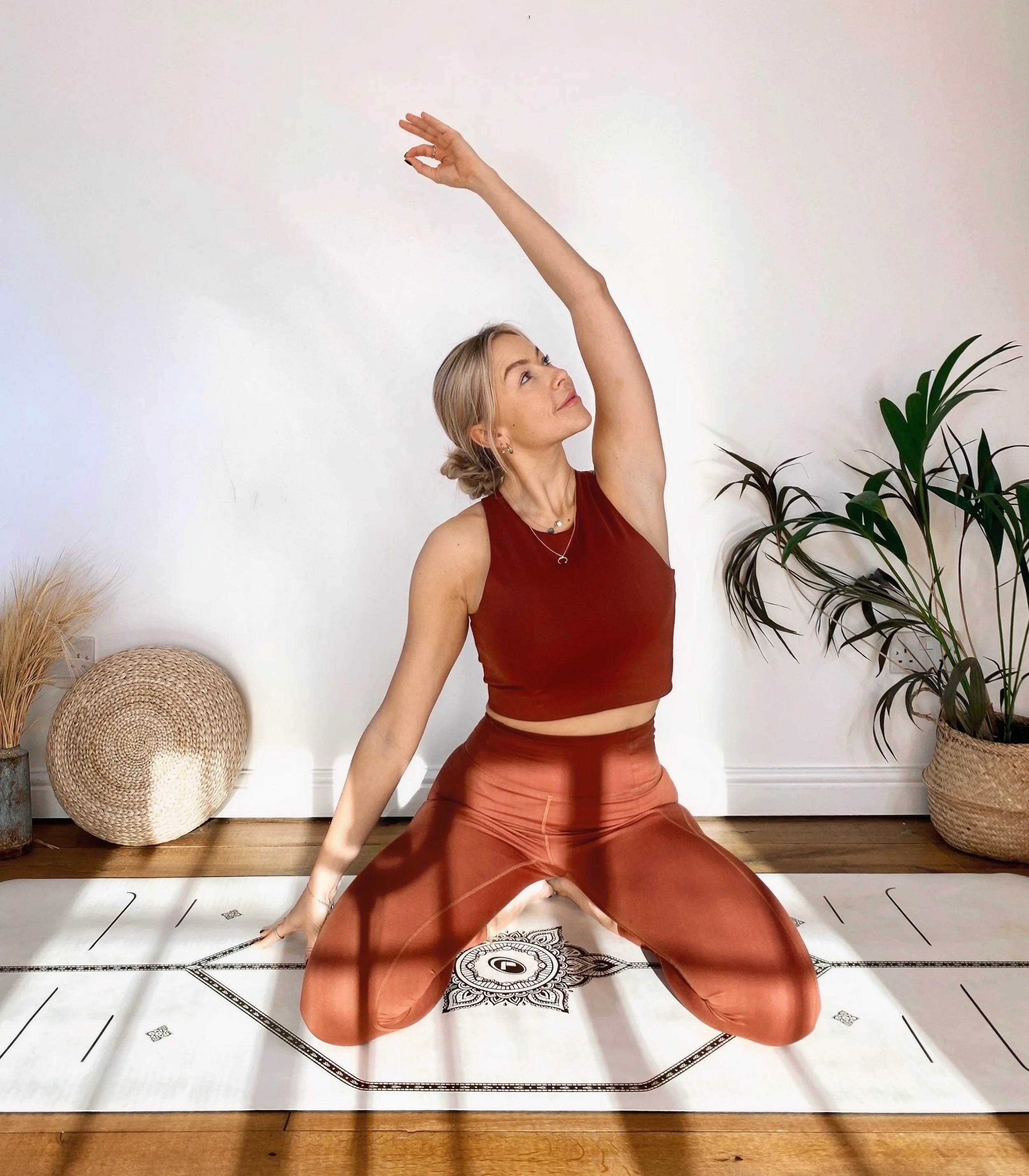 Abbi our yoga teaching doing yoga pose indoors on a mat, wearing red workout clothes, with plants and wicker basket nearby.