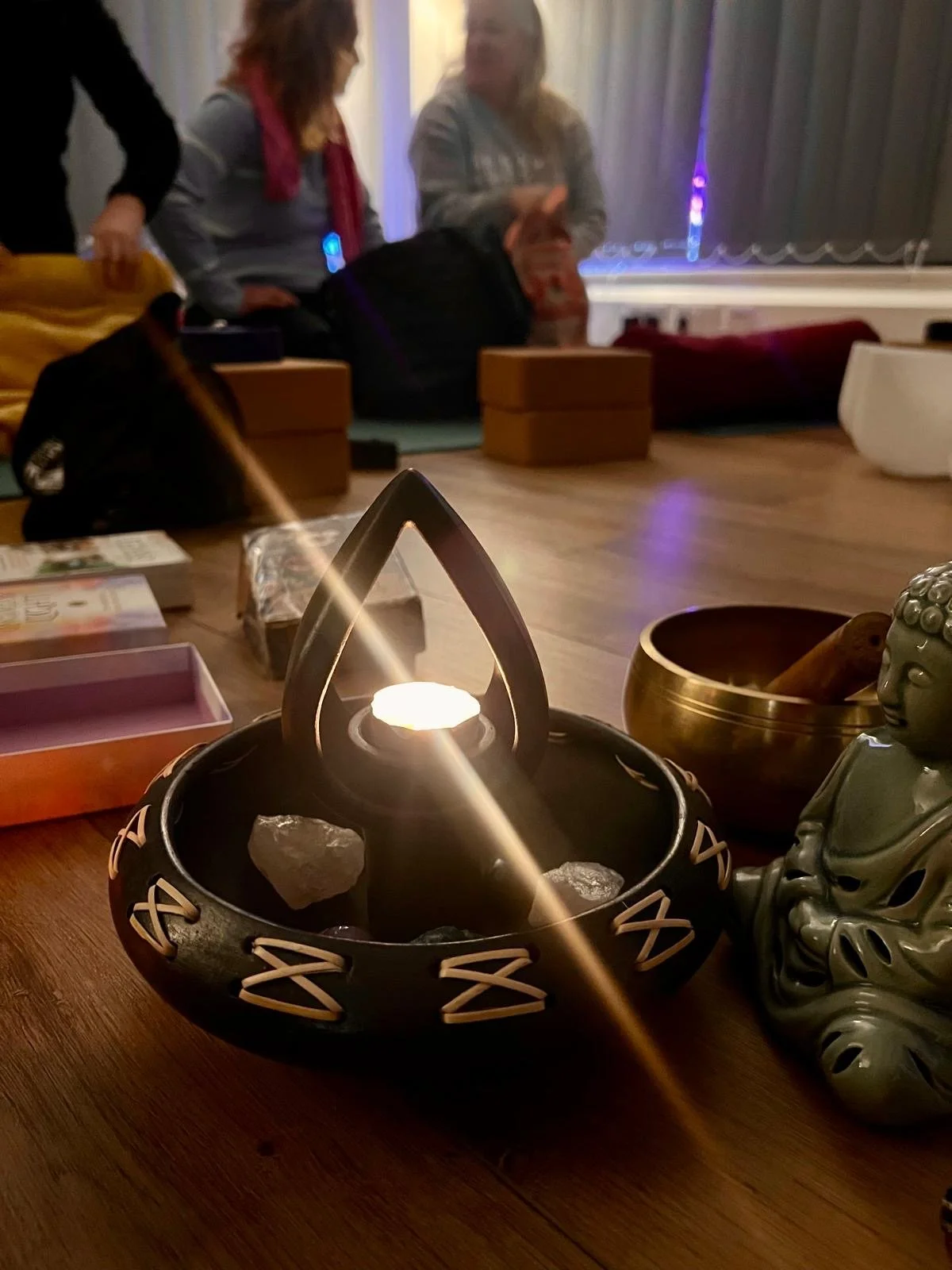Flourish Yoga Studio in Teddington. A dimly lit room with a central candle holder surrounded by crystals, a Buddha statue, and meditation accessories. People are sitting on the floor in the background with meditation cushions and blocks.