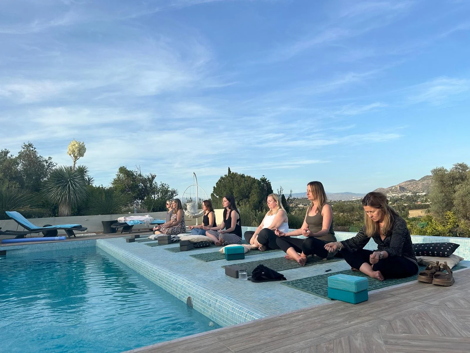 Group of people meditating by a swimming pool in an outdoor setting with trees and a mountainous background.