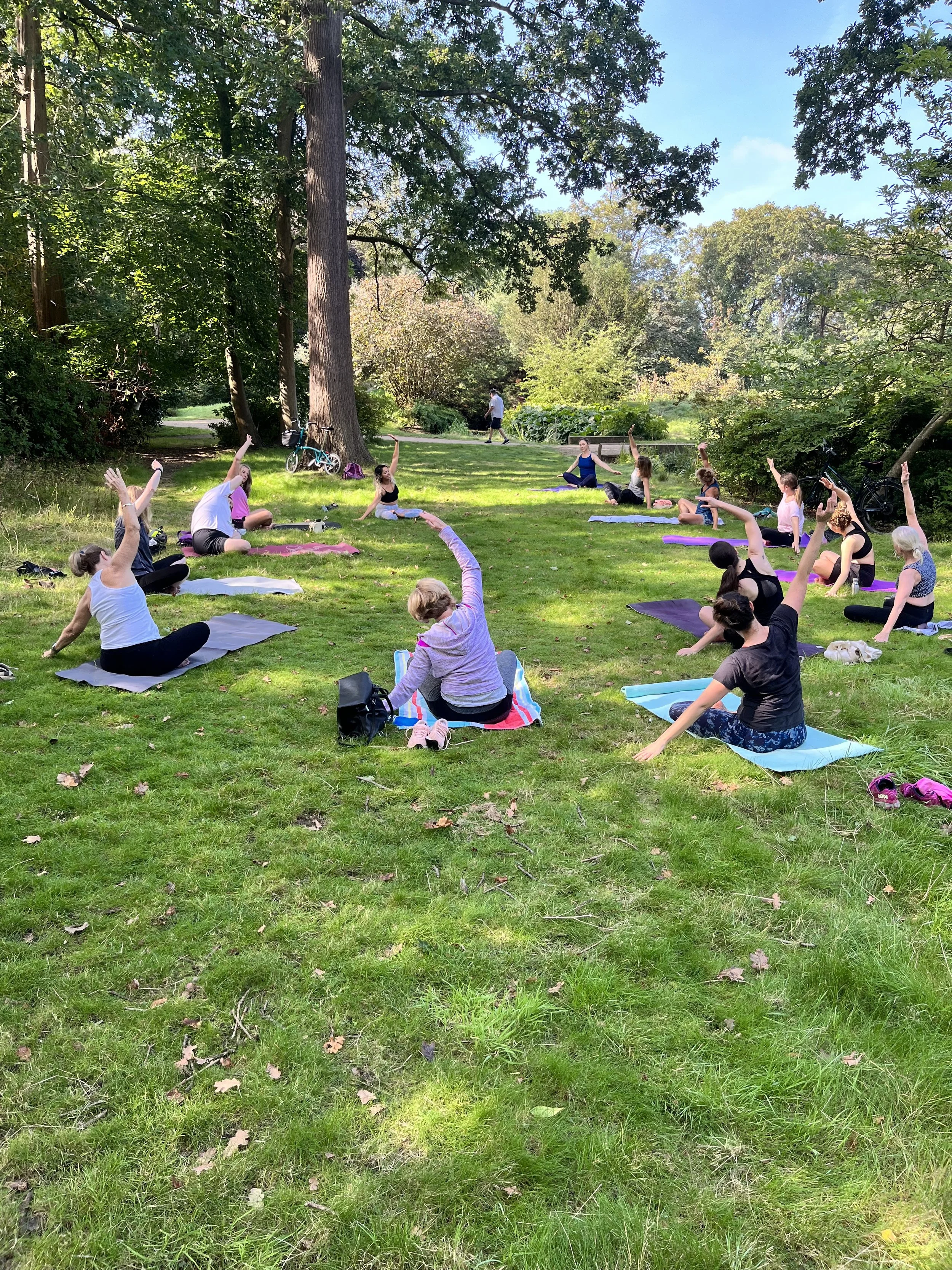 Group yoga session in a park with people sitting on mats, stretching, surrounded by lush greenery and trees.
