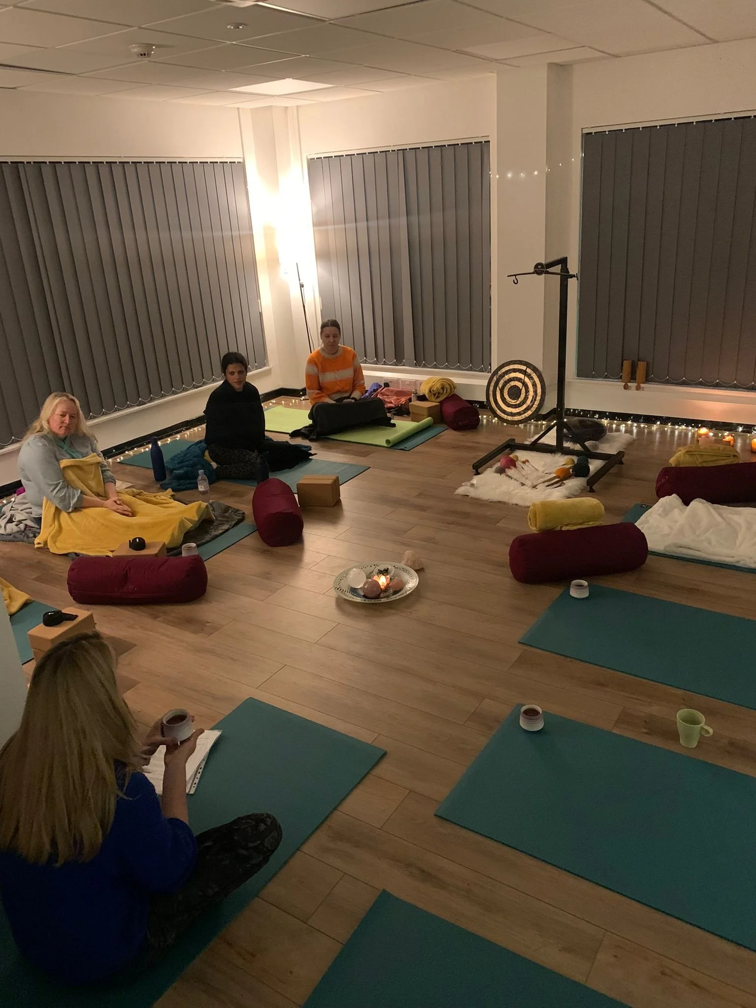 A group of people sitting on yoga mats in a dimly lit room. The room has soft lighting, yoga props like bolsters and blocks, and a circle of candles in the center. Participants are seated comfortably, possibly in a meditative or relaxed setting.