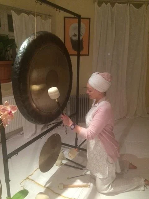 Person in white attire playing a large gong in a serene room with curtains and a plant.