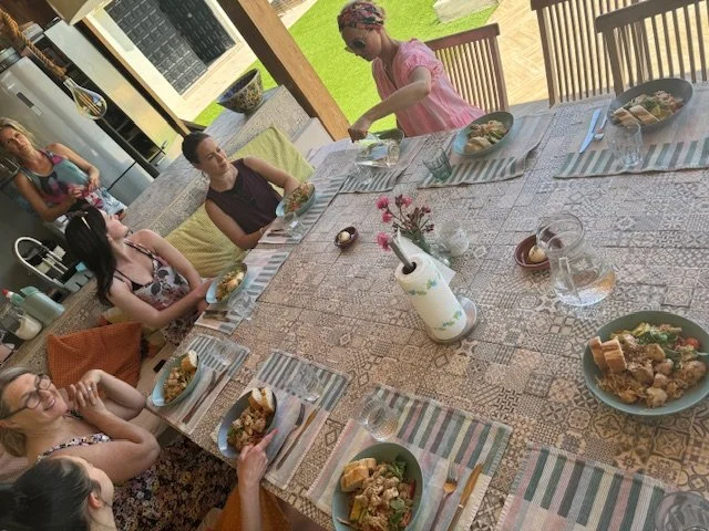 A group of people sit around a dining table set for a meal, with dishes of food and drinks on patterned placemats. The setting appears to be an outdoor patio with a view of grass and garden area. One person is standing, while the rest are seated, eng