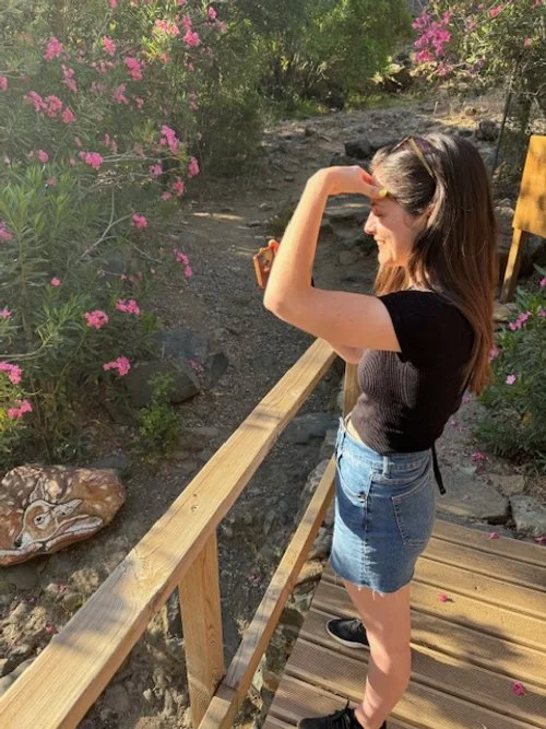 A young woman in a black top and denim skirt standing on a wooden bridge, looking out into the distance while shielding her eyes from the sun. She is surrounded by pink flowering bushes and trees in a natural setting.