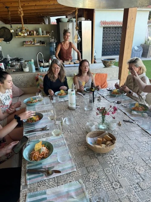 Group of people sitting at a dining table outdoors, eating meals in colorful bowls, with a woman in the background near a kitchen area.