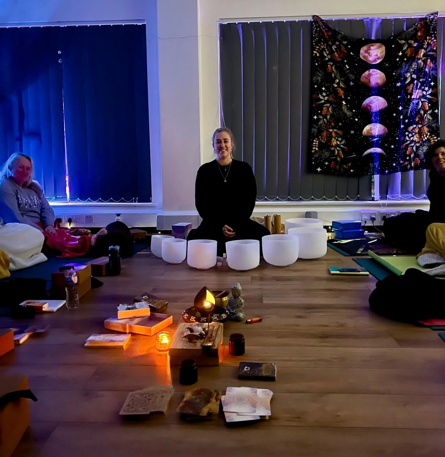 A dimly lit room with a group of people sitting on the floor. In the center, there are various objects like candles, books, and decorative items arranged on a wooden floor. One woman is sitting facing forward with several white bowls, possibly crystal singing bowls, in front of her. The room has vertical blinds and a decorative tapestry with moon phases hanging on the wall.