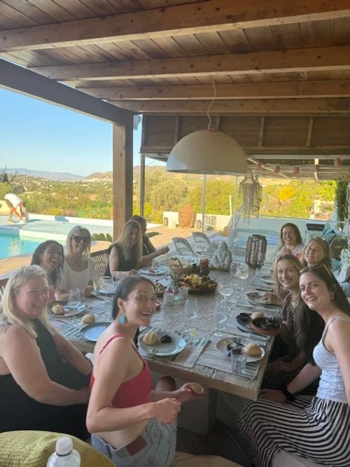 Group of women at a dining table by a pool in an outdoor setting under a wooden pergola.