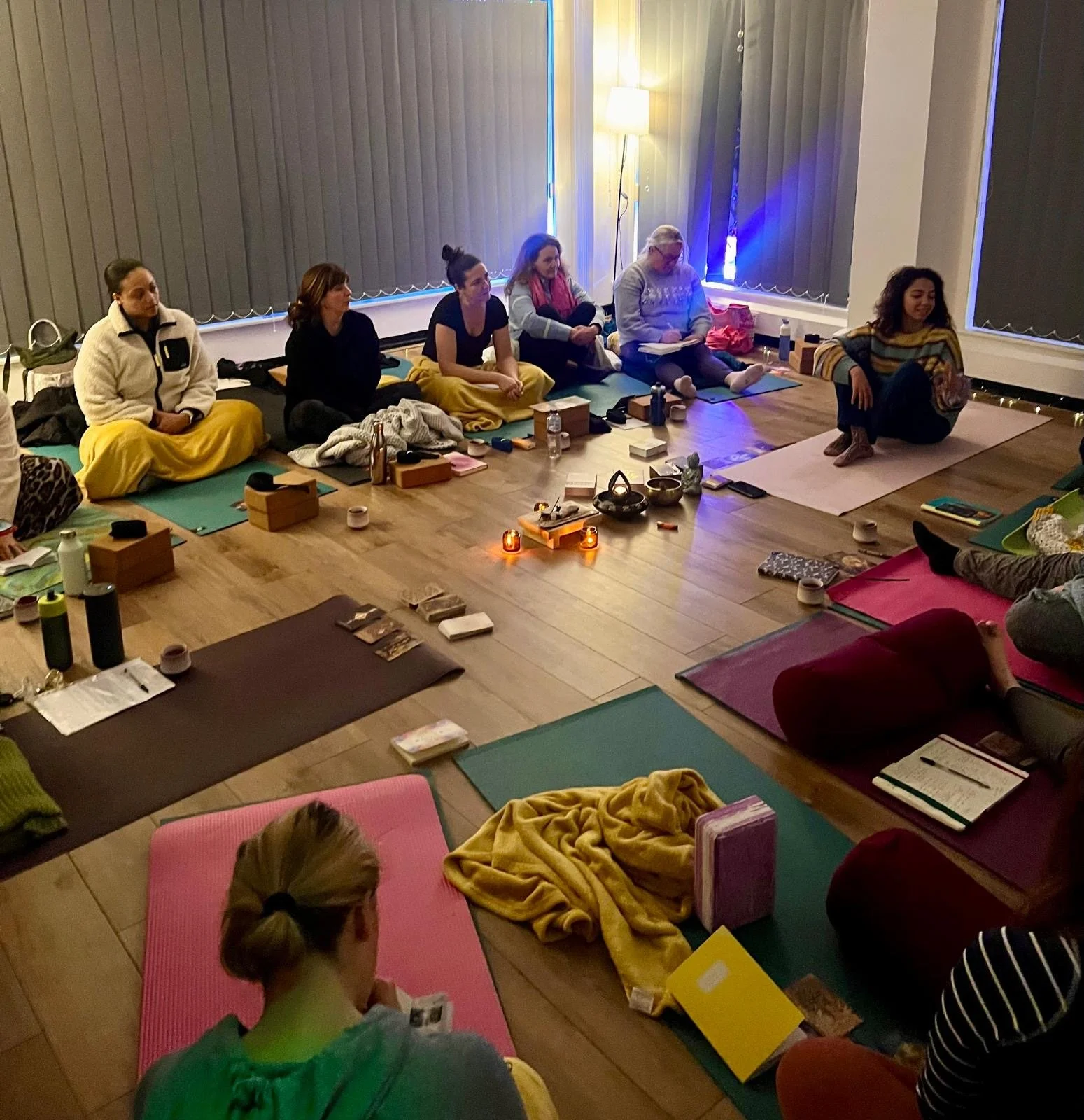 A group of people sitting in a circle on yoga mats in a dimly lit room, surrounded by candles and personal items, suggesting a meditation or wellness session.