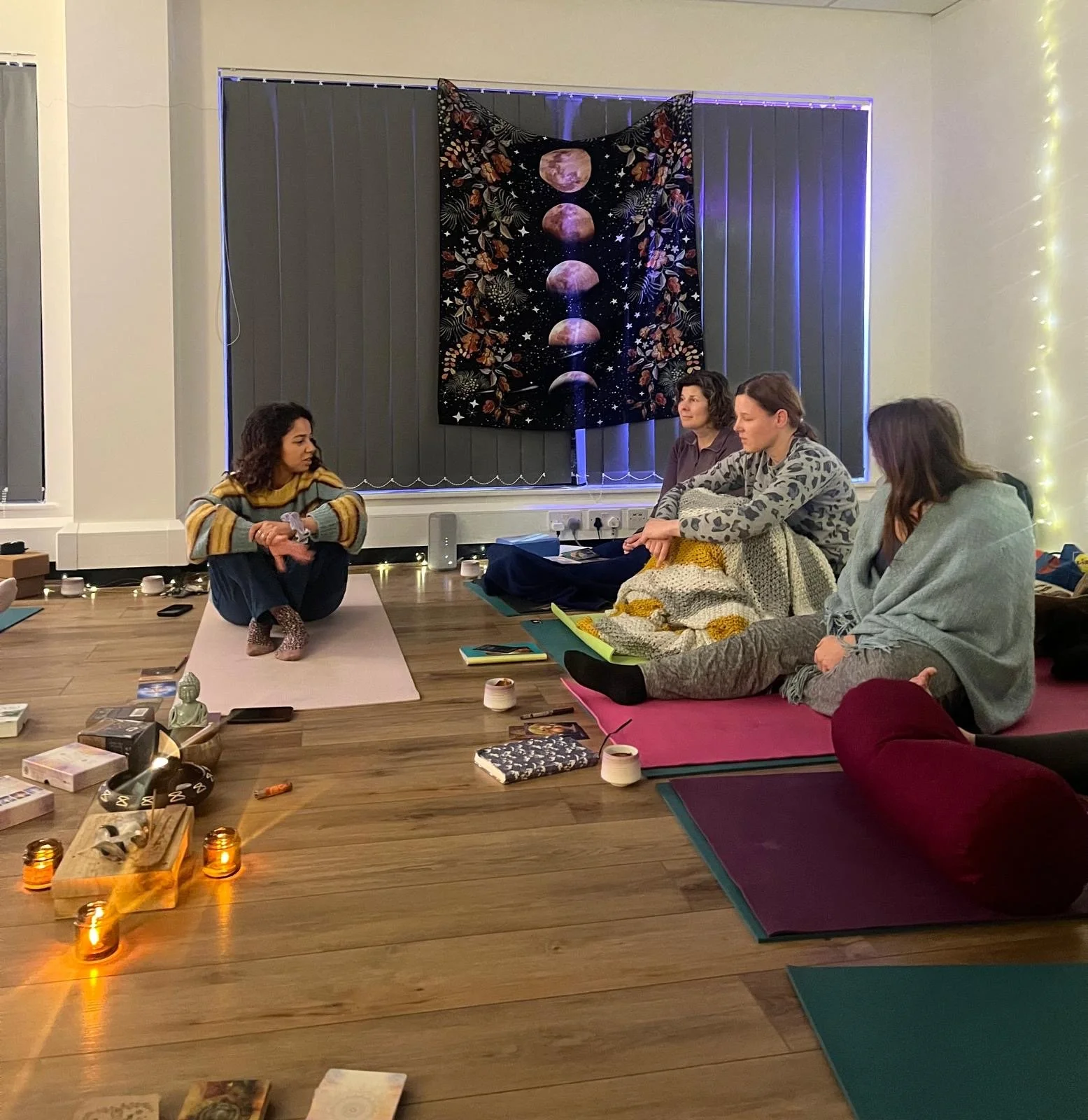 A Flourish yoga moon workshop - a group of people sitting on yoga mats in a dimly lit room, with blankets, candles, and cushions, creating a cozy, spiritual atmosphere. A tapestry with moon phases hangs on the wall.
