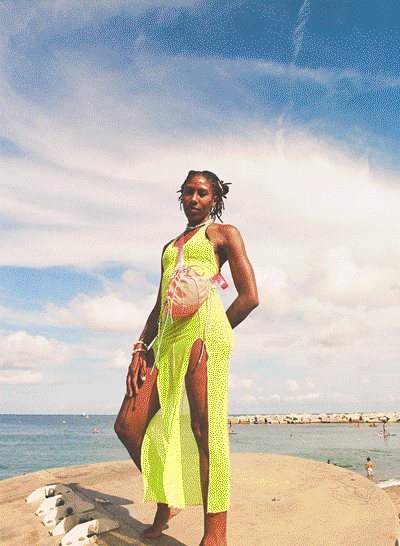Woman standing on a beach, wearing a neon green dress, with a bag over her shoulder, under a partly cloudy sky.