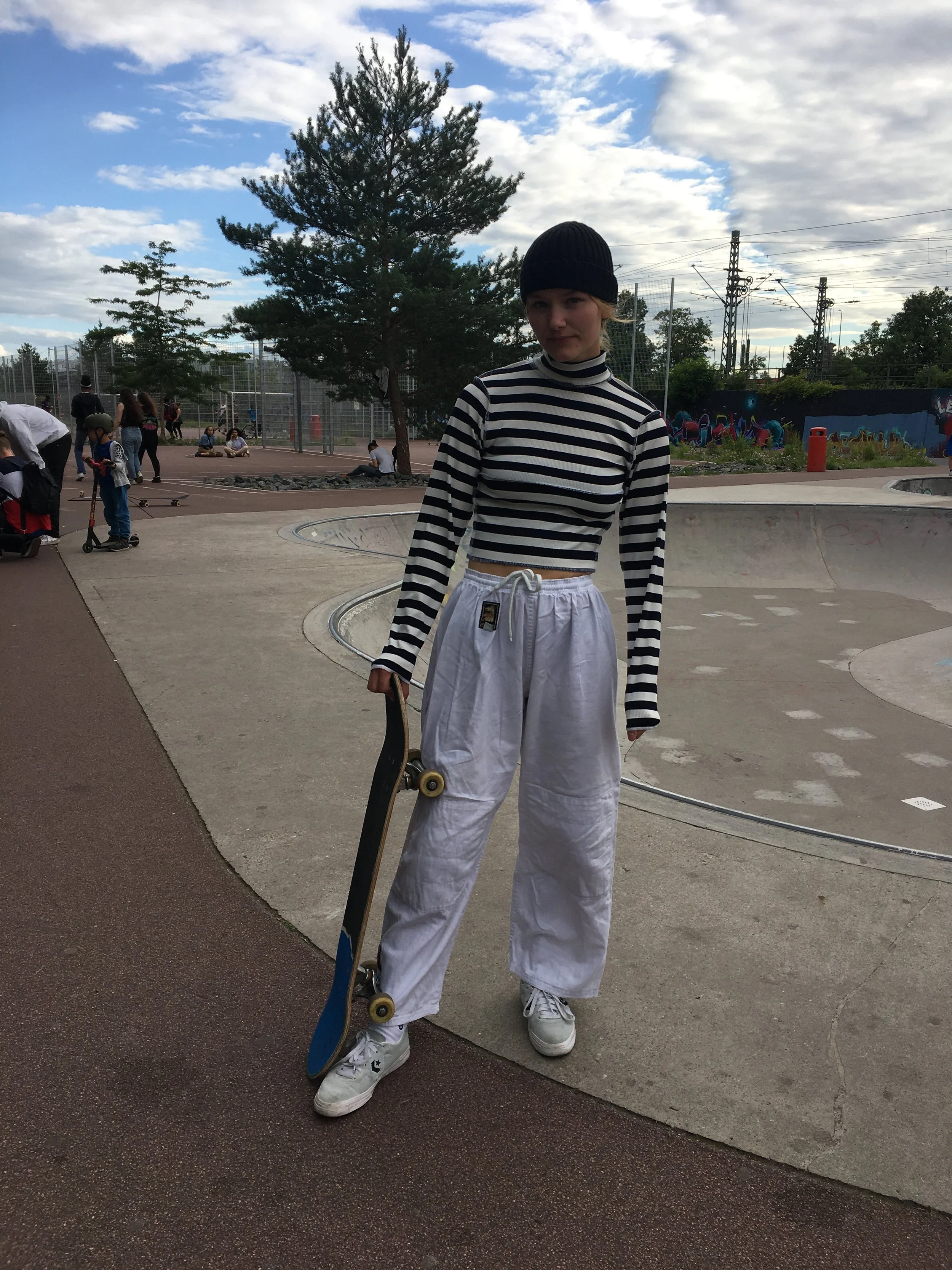 A young woman in a black beanie, black and white striped long sleeve crop top, white baggy pants, and white sneakers, holding a skateboard, standing at a skatepark with other skaters, trees, and a graffiti wall in the background under a partly cloudy