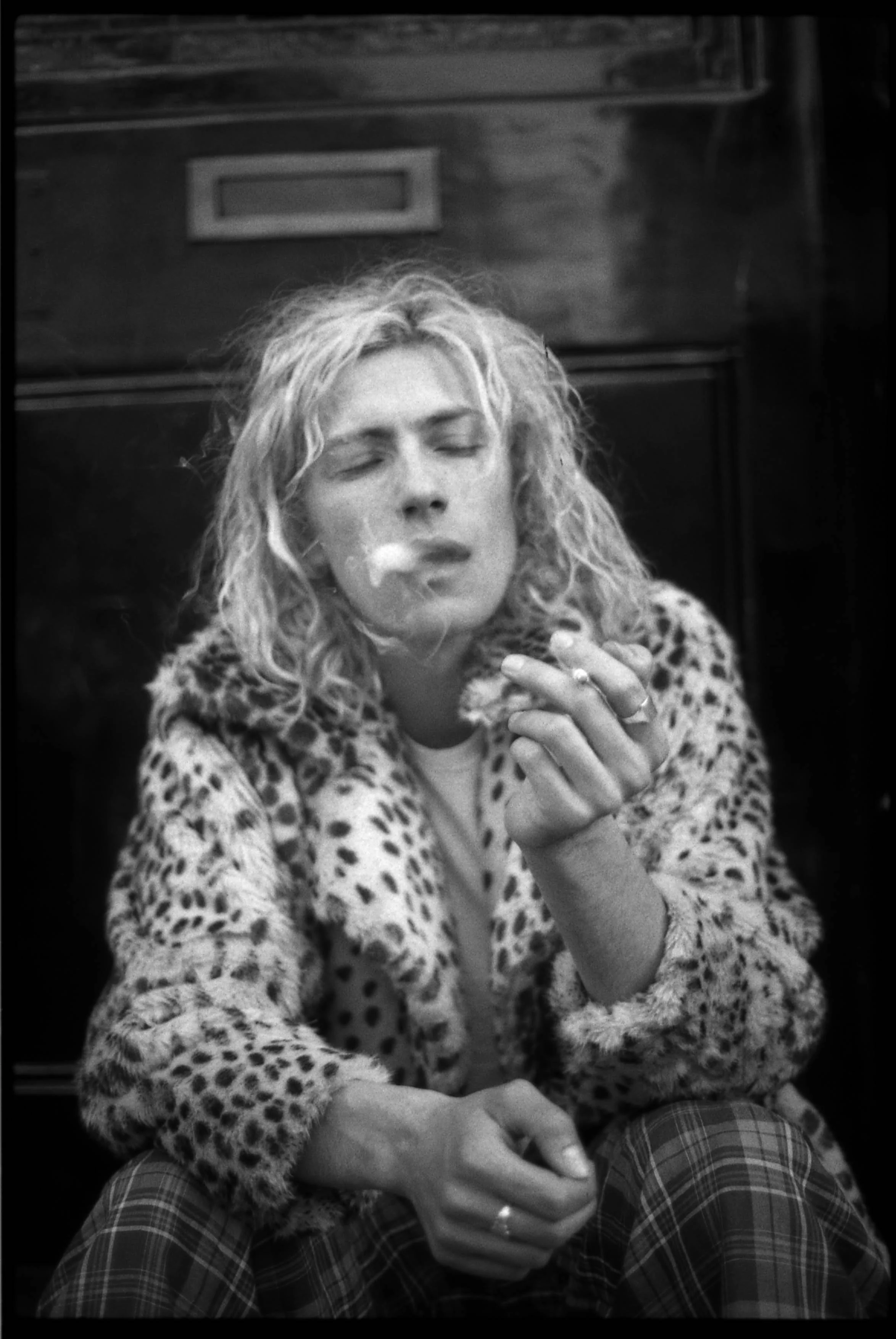 A woman with curly blonde hair, wearing a leopard print coat and checkered pants, smokes a cigarette with her eyes closed.