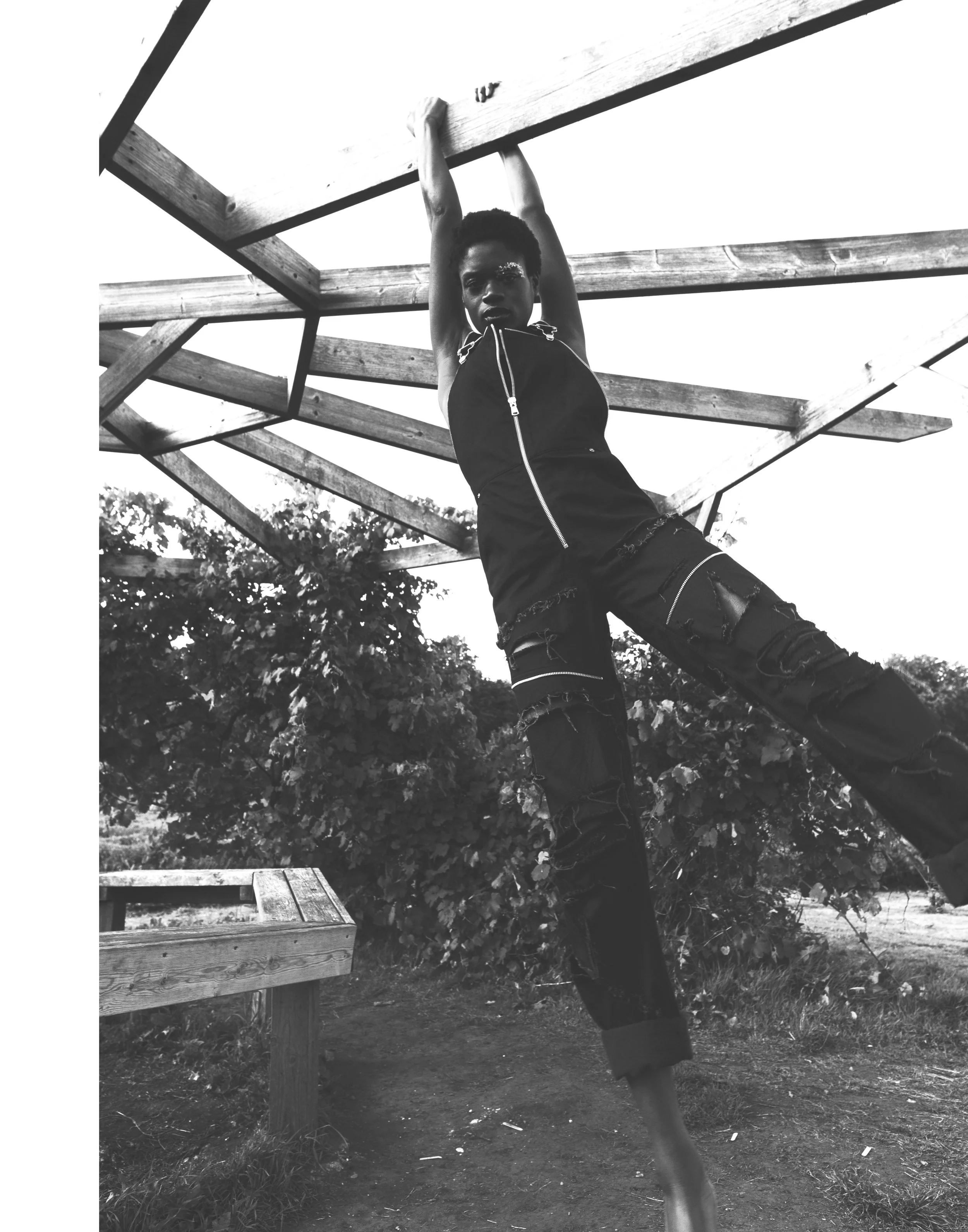 A woman hanging from a wooden structure outdoors, wearing a sleeveless zip-up top and ripped Jeans.