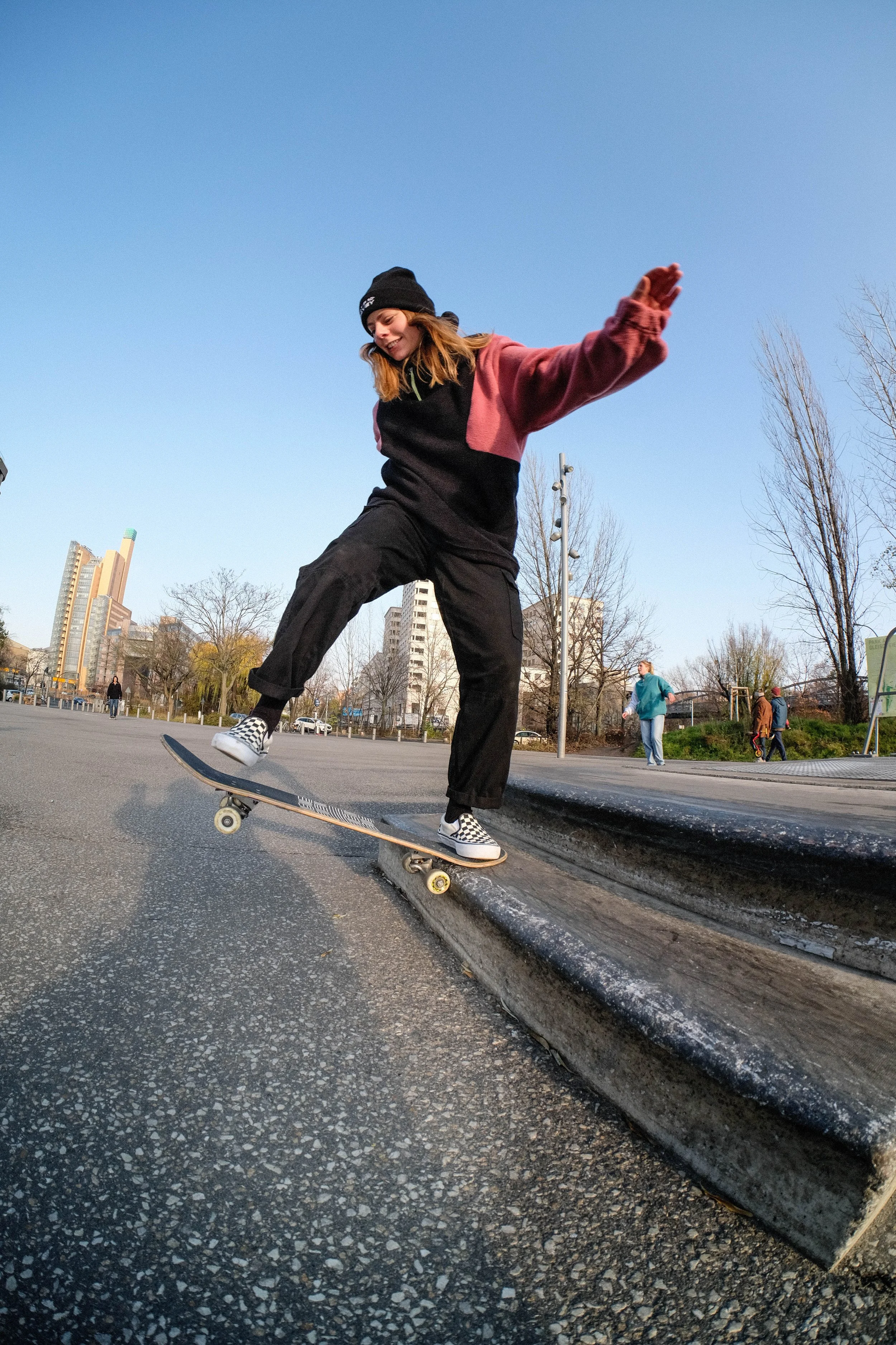 A young woman skateboarding on a curb in a park with other people in the background, city buildings, and trees under a clear blue sky.