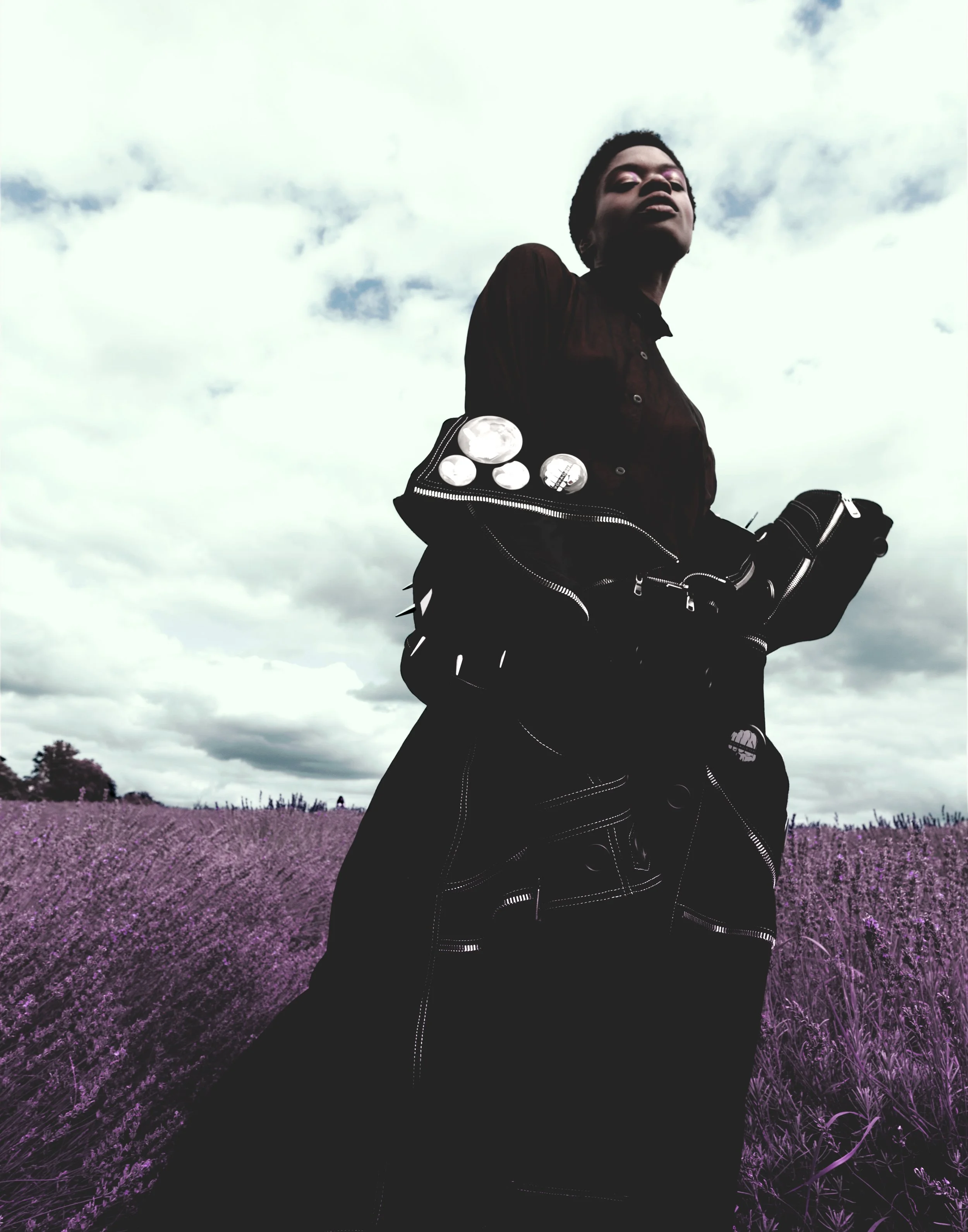 Person standing in a field of purple flowers with a cloudy sky background, wearing a dark outfit and carrying a bag