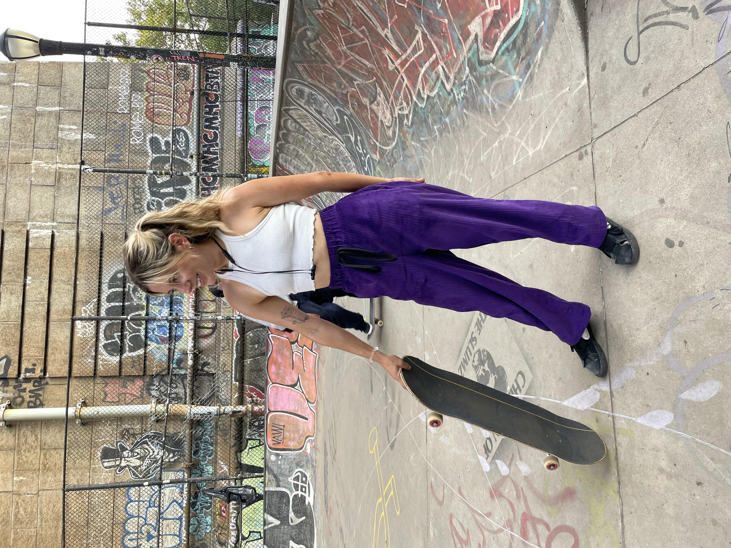 A young woman with long blonde hair, wearing a white tank top, purple pants, and black sneakers, holding a skateboard, standing on a graffiti-covered skate park floor near a chain-link fence.