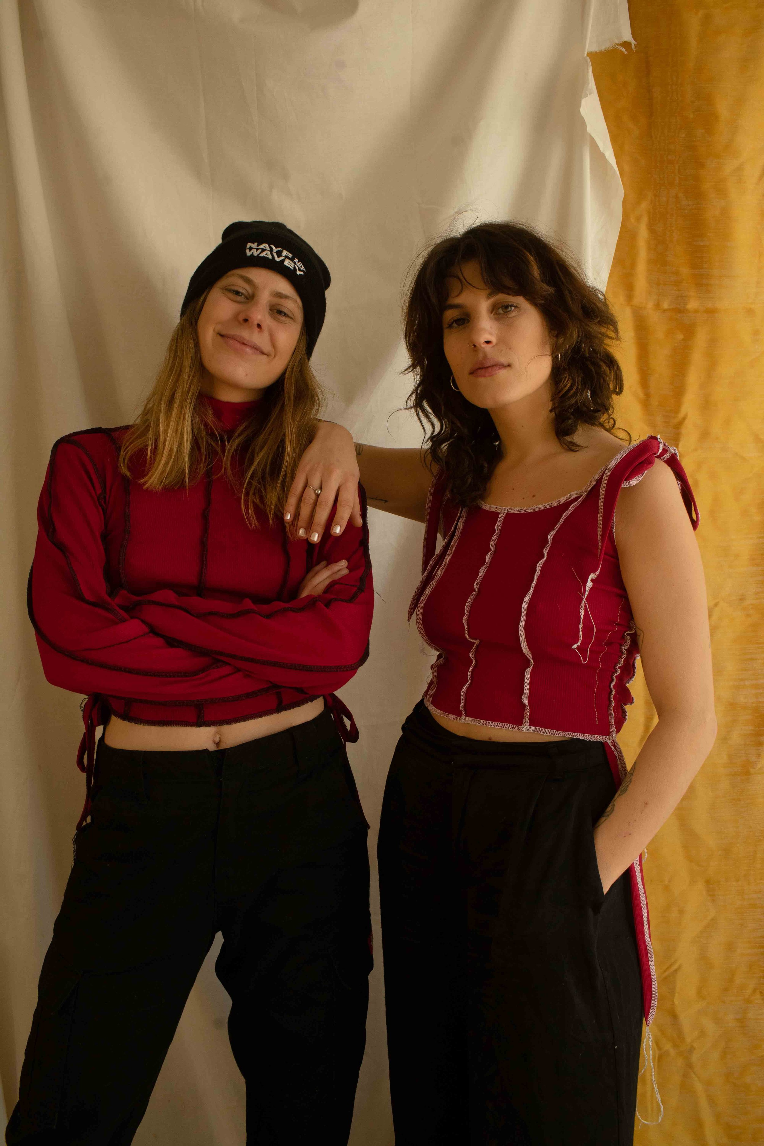 Two young women standing side by side, one with arms crossed and the other with her hand in her pocket, in front of a beige and yellow backdrop.