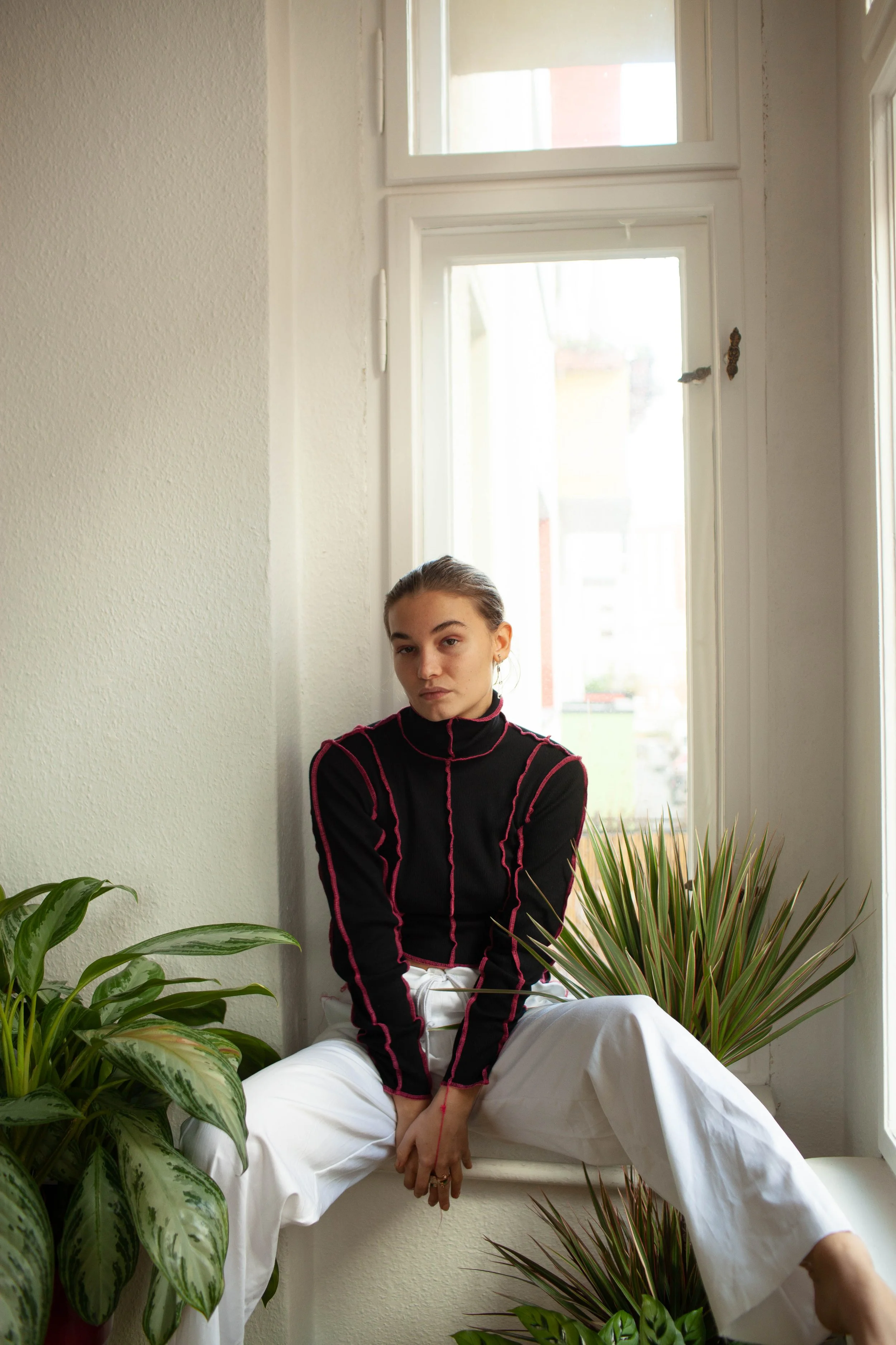 A young woman sitting on a windowsill, surrounded by green plants, wearing a black turtleneck with pink piping and white pants, indoor setting with natural light.