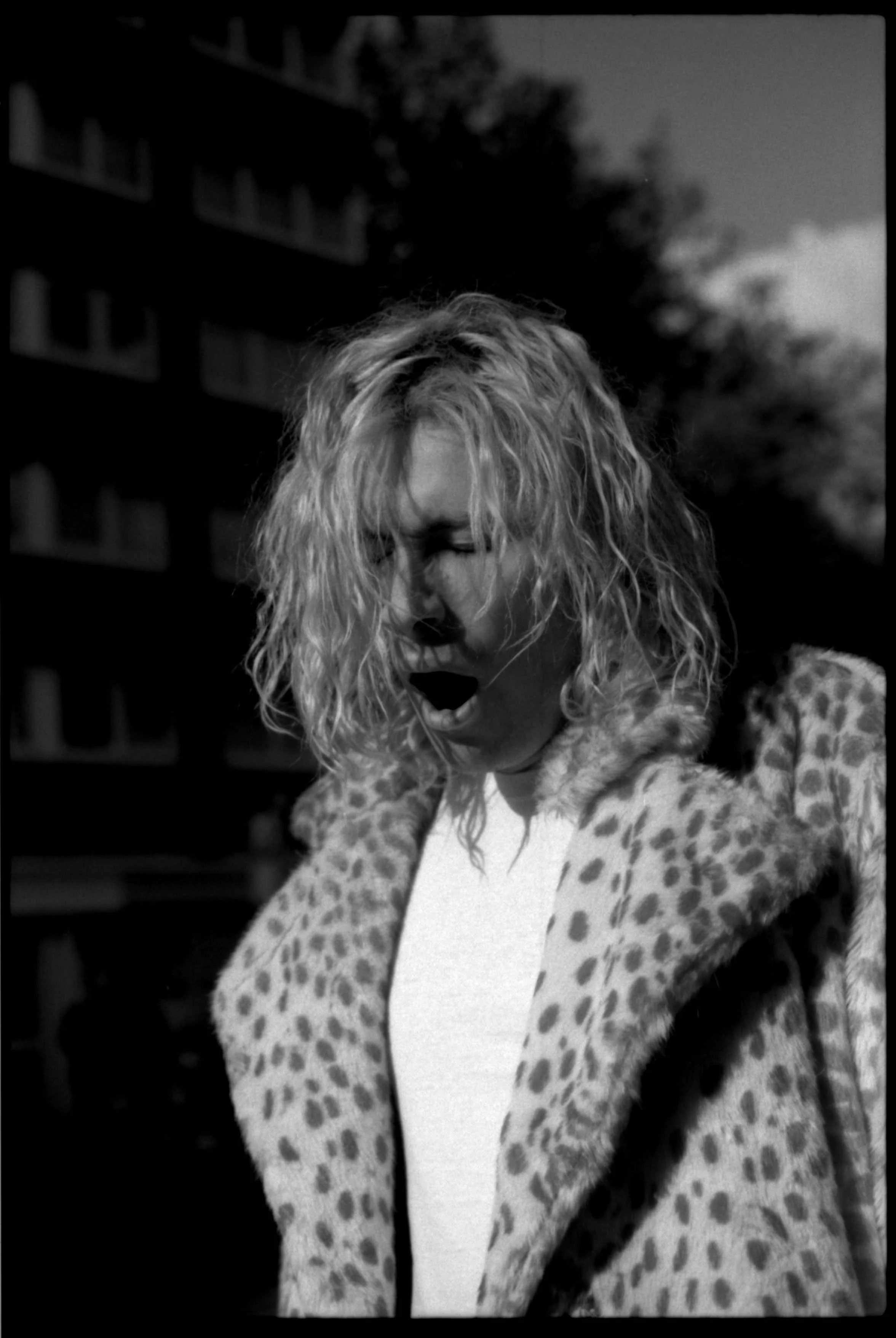 A black and white photo of a woman with curly, shoulder-length hair singing or crying with her eyes closed. She is wearing a leopard print coat and a light shirt, outdoors with a blurred building and trees in the background.