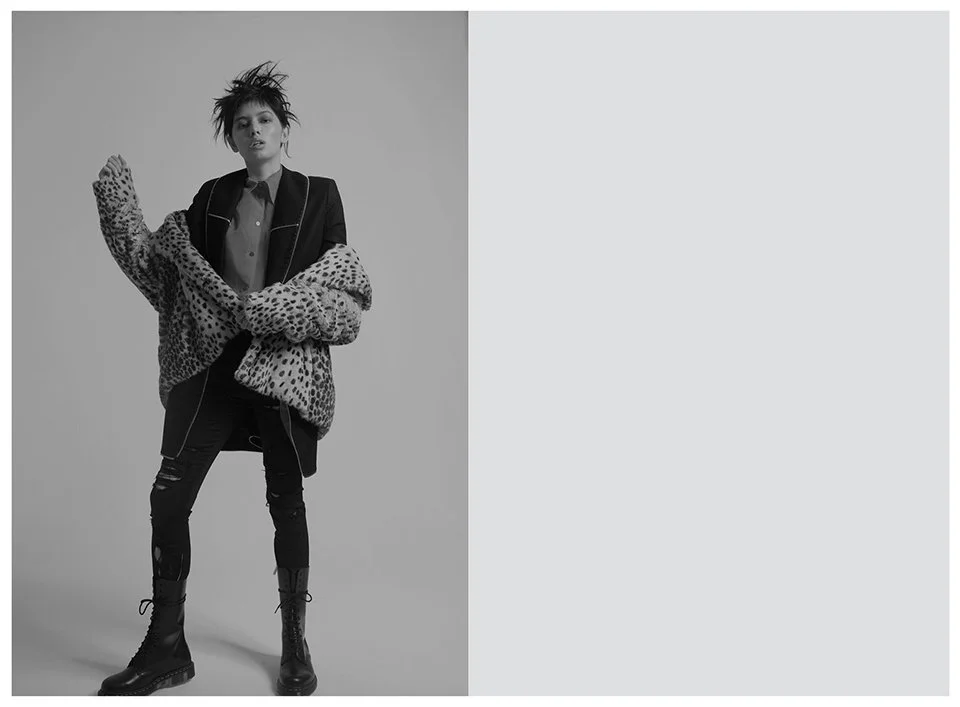 A black and white photo of a young woman with short, spiky hair, wearing a leopard print coat, black pants, and combat boots, standing against a plain background.