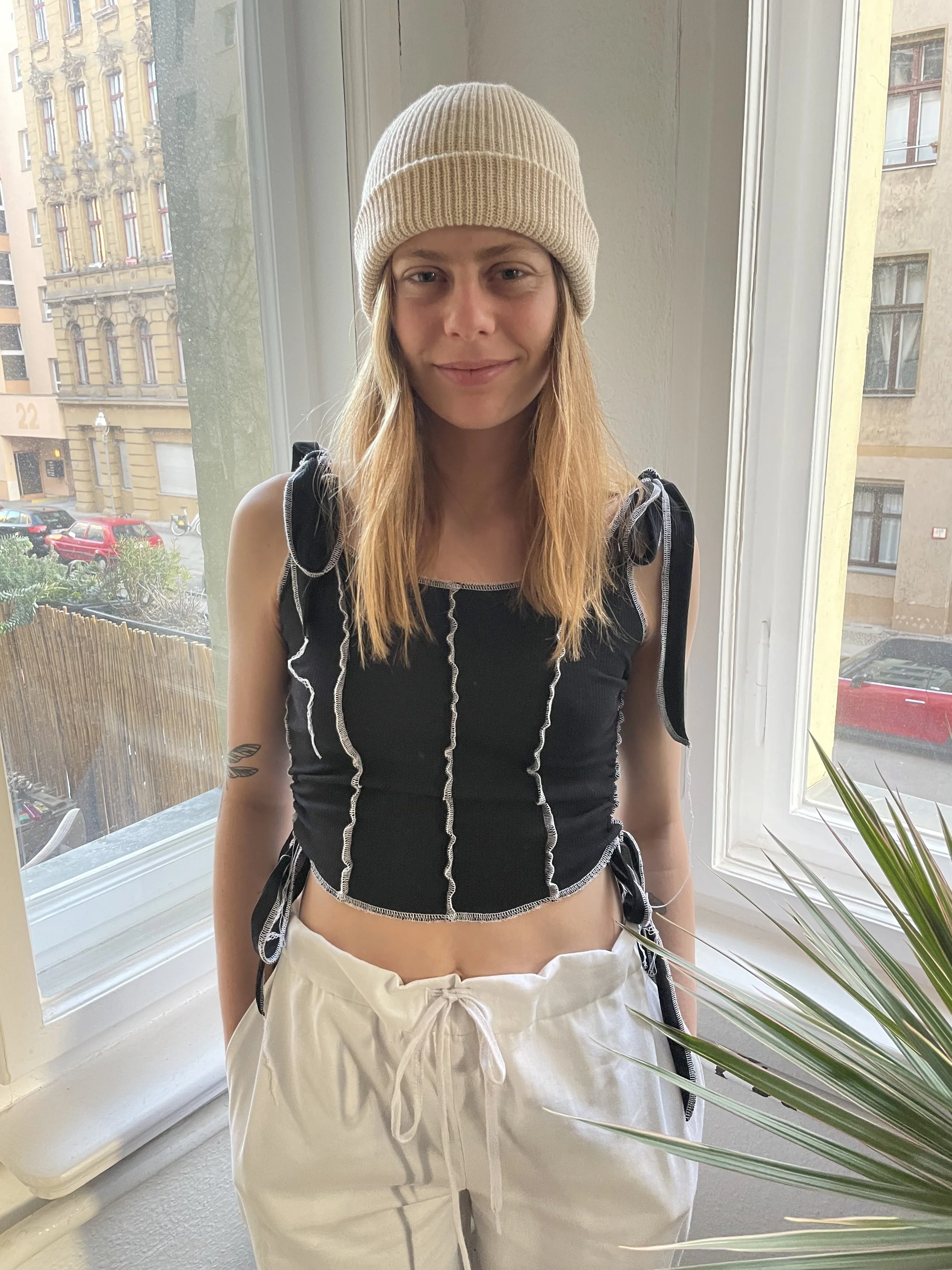A young woman with long red hair wearing a beige knit beanie, a black top with white stitching and tied shoulder straps, and white drawstring pants standing by a window.