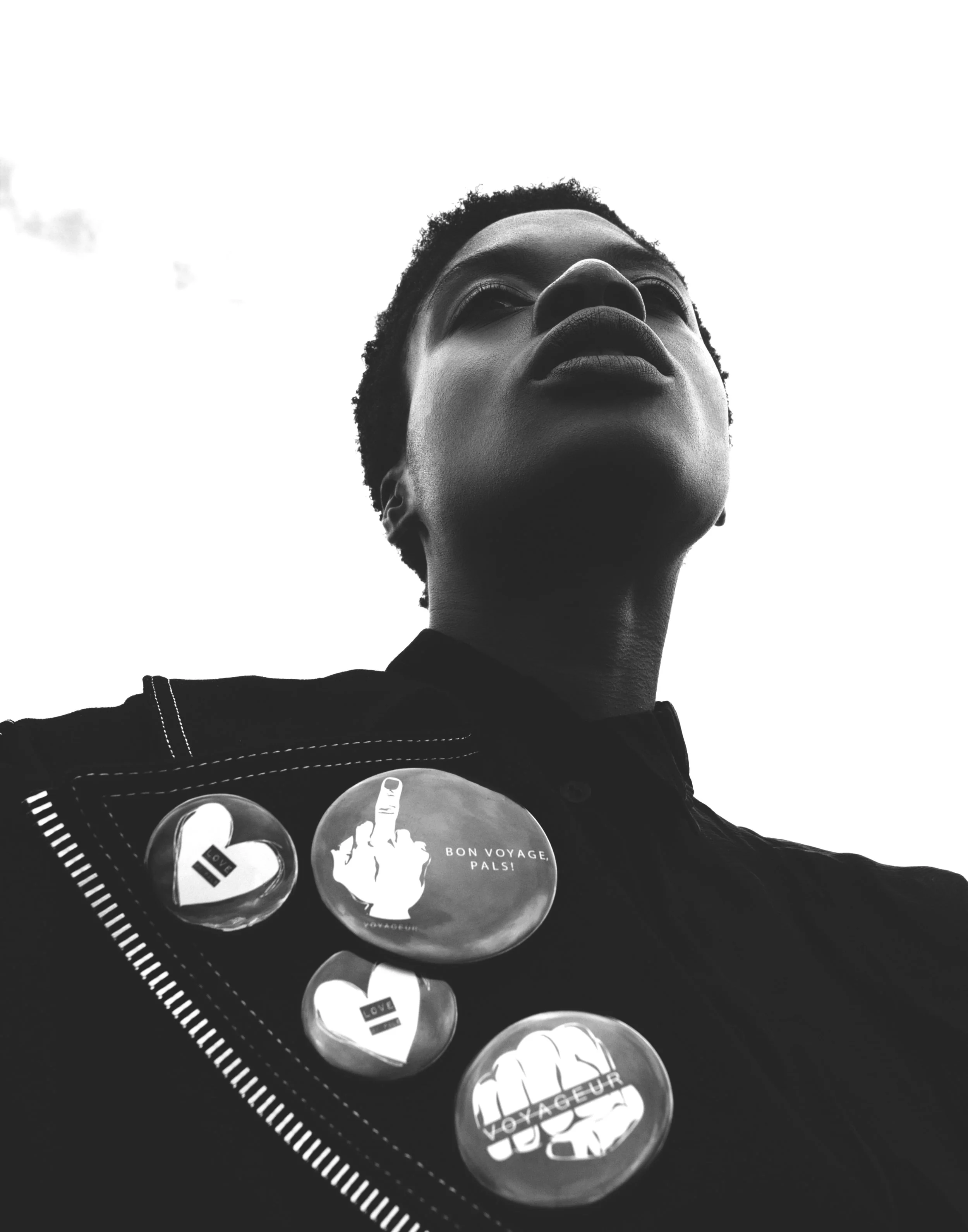 A black and white photo of a woman looking upward, with short natural hair, wearing a jacket decorated with four pins, two of which have a raised middle finger and messages like "BON VOYAGE. PALS" and "VOYAGEUR."