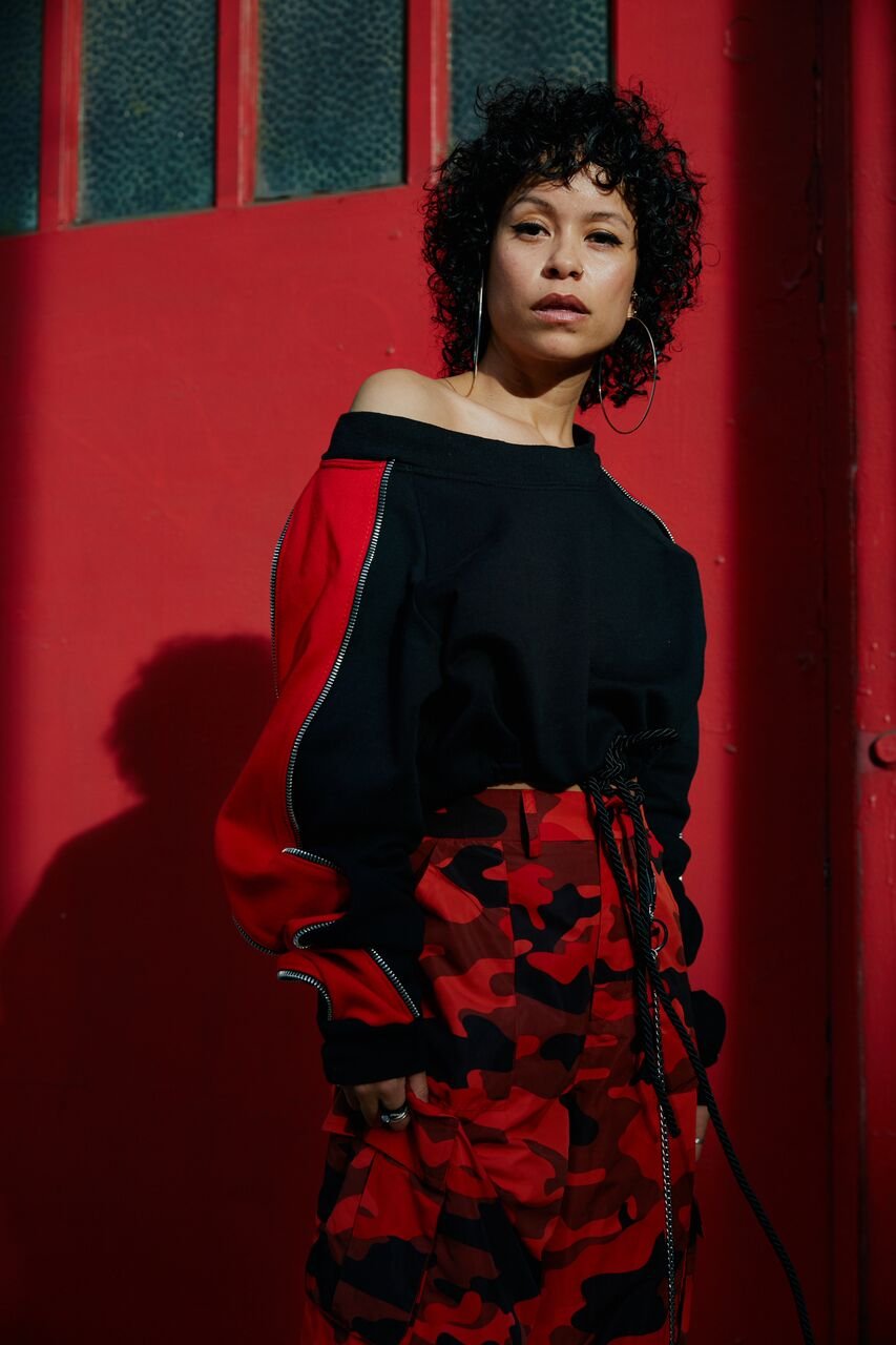 A woman with curly black hair, wearing a black off-the-shoulder sweatshirt and red camouflage pants, standing against a red background.