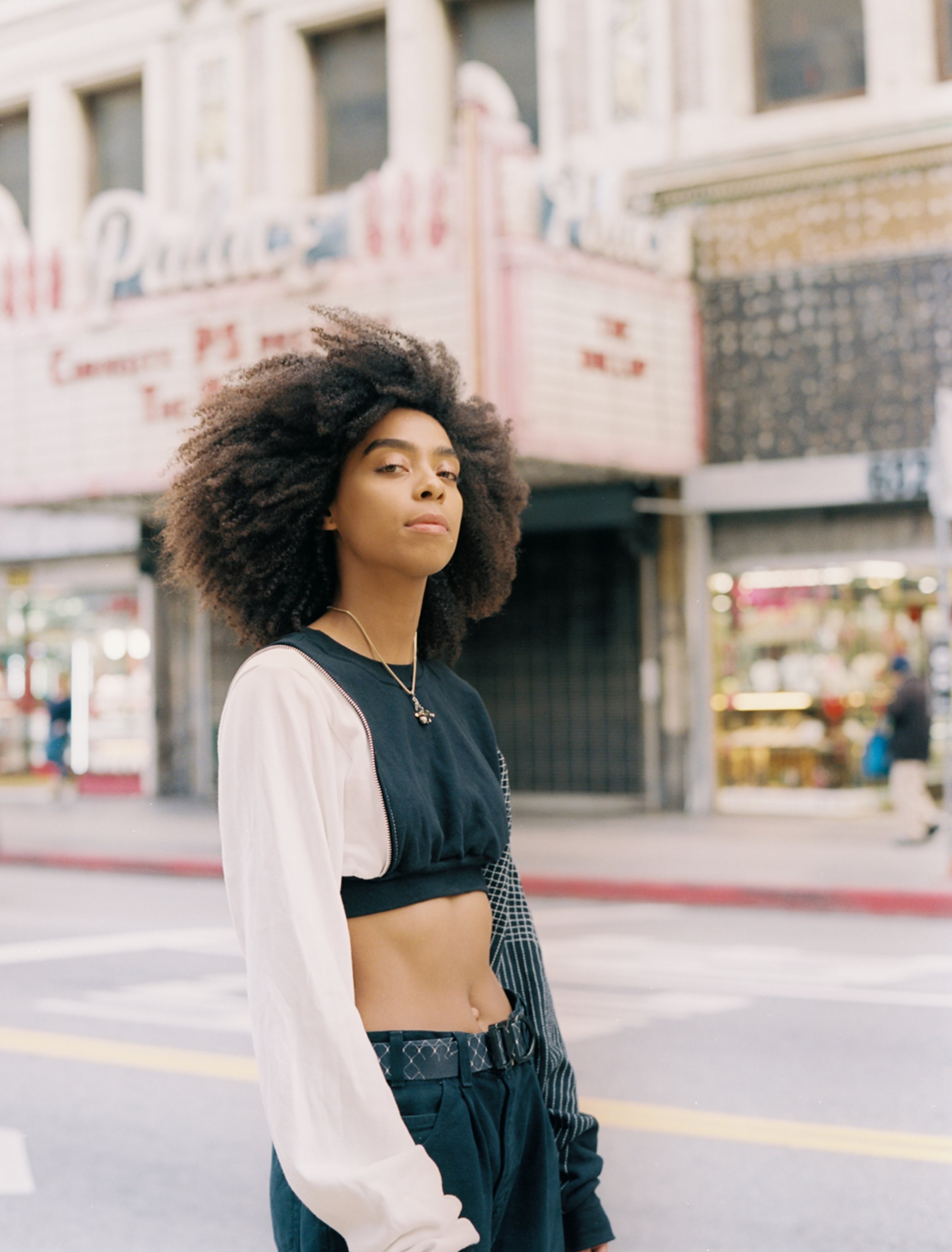 Young woman with voluminous curly hair standing on a city street, wearing a black and white crop top, black pants, and layered necklaces, with a vintage theater in the background.