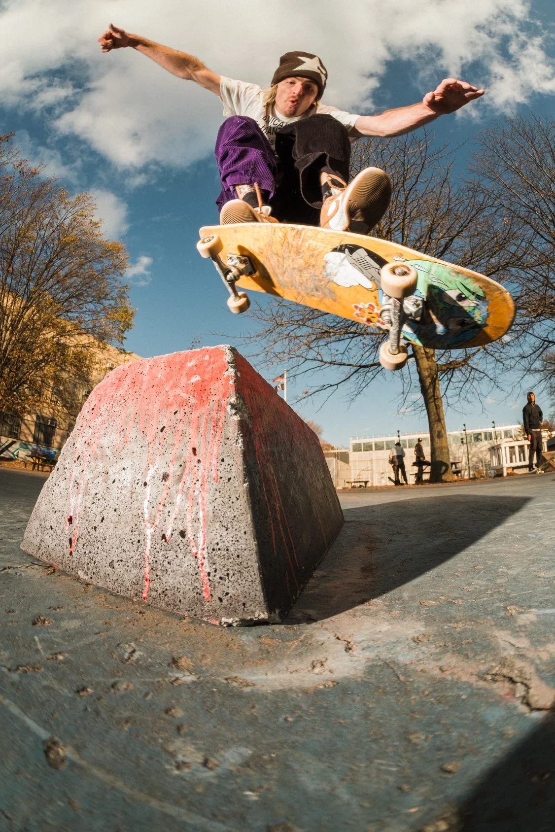Skateboarder in mid-air performing a trick over a concrete obstacle in an outdoor skate park with a blue sky and leafless trees in the background.