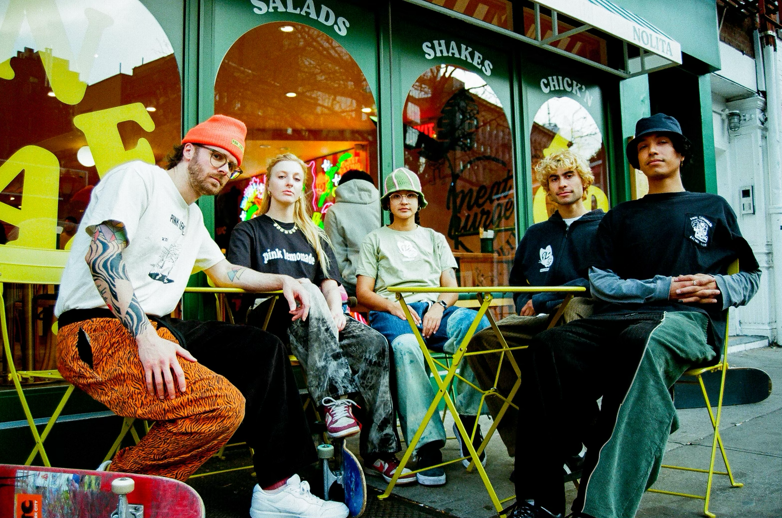 Group of five young people sitting outside a colorful restaurant with neon signs, some with skateboards, dressed in casual streetwear.