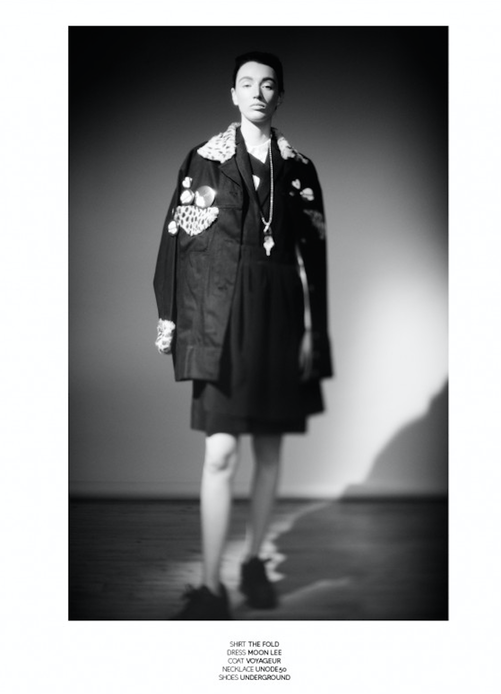Black and white photo of a female model standing indoors against a plain wall, wearing a dark jacket with colorful patches, a dress, a coat, a necklace, and shoes.