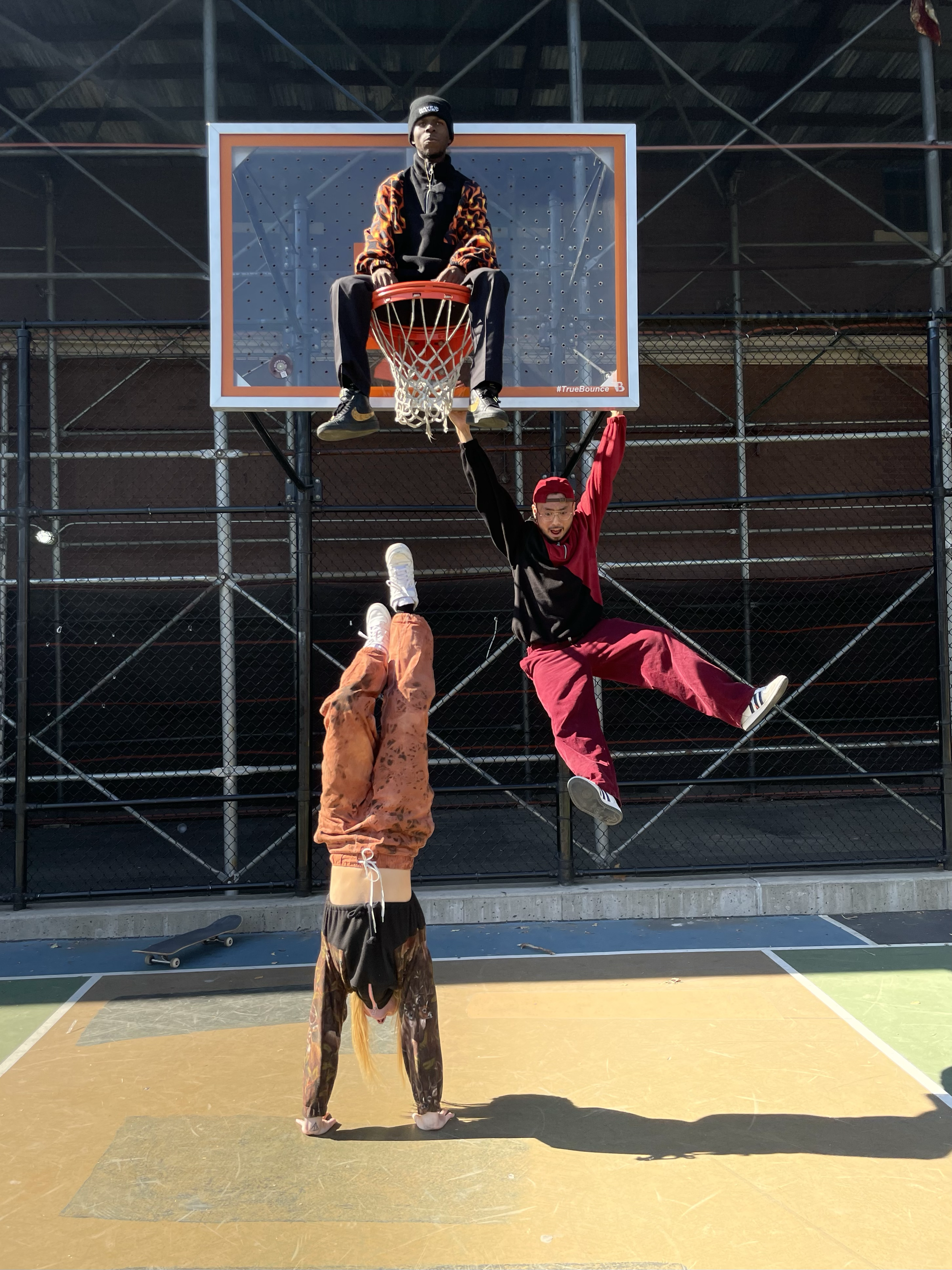 Three people on an outdoor basketball court, with one person doing a handstand and two others hanging from the basketball hoop, all appearing to be having fun.